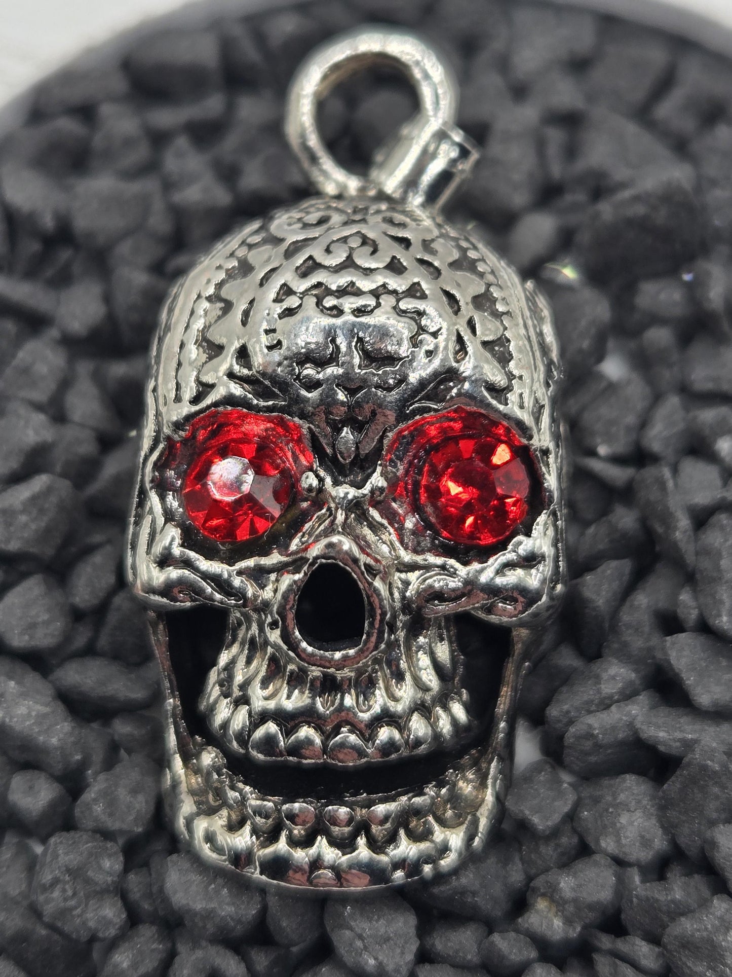 A silver skull pendant with red gemstones is shown against a dark background of small rocks.