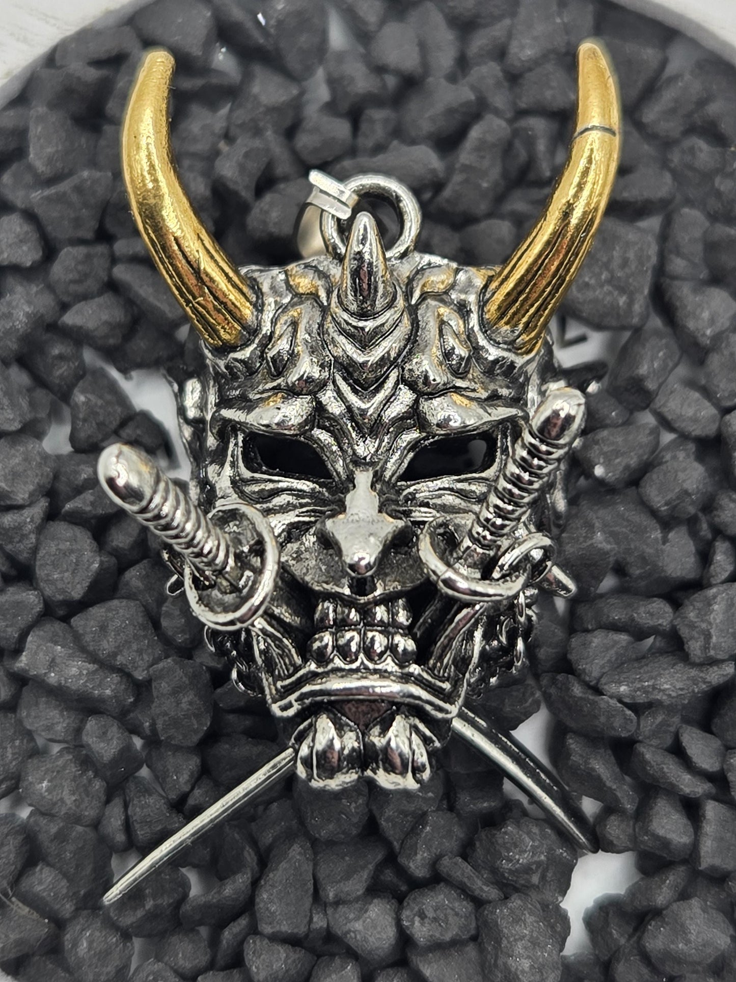 A detailed metal sculpture of a skull with horns and a sword, set against a background of small rocks.