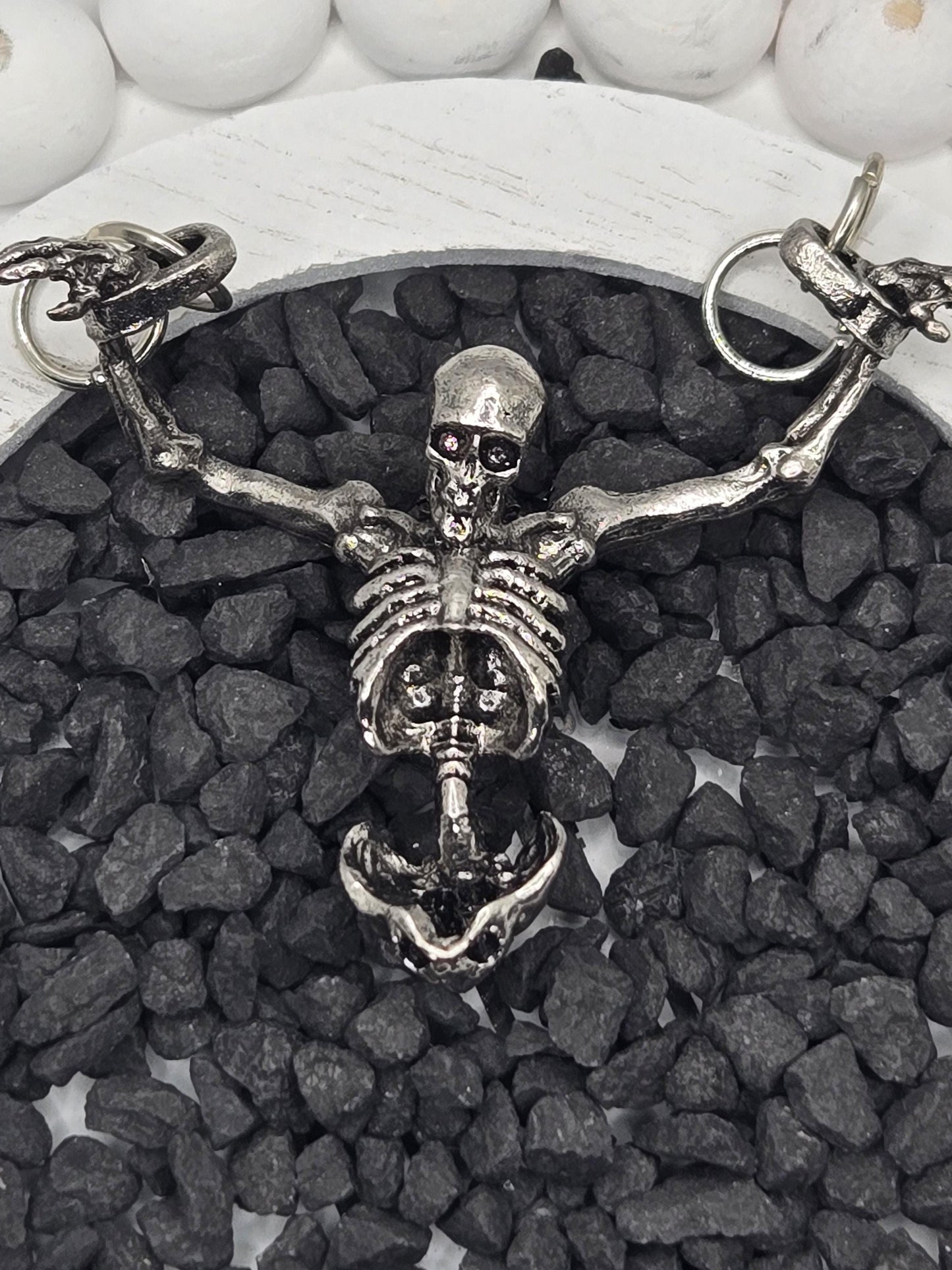 A silver skeleton necklace with a pendant is displayed on a bed of small black stones.