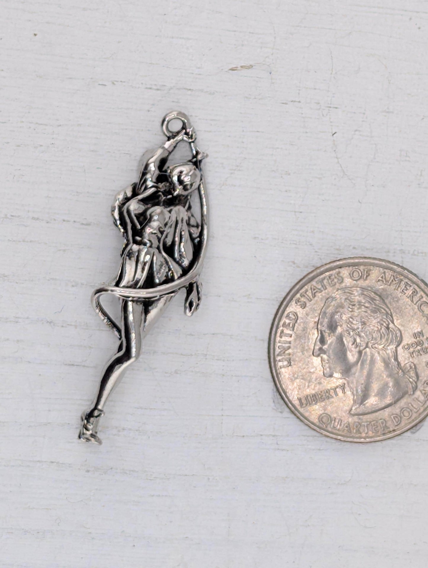 A silver-colored metal charm in the shape of a woman is placed next to a United States quarter coin on a white surface.