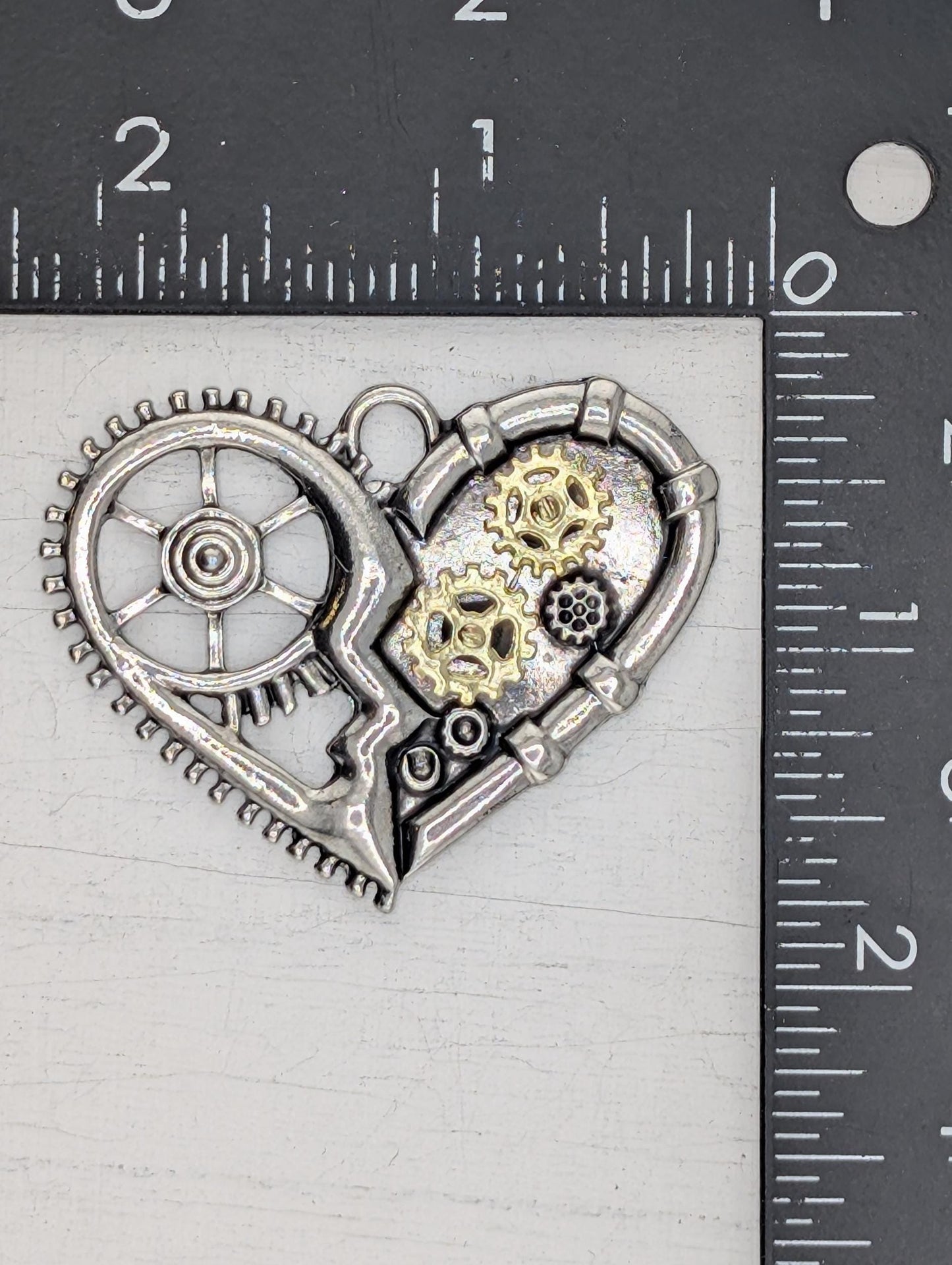 A close-up view of a silver-colored metal heart-shaped pendant with gears and cogs attached to it, placed on a ruler for size comparison.