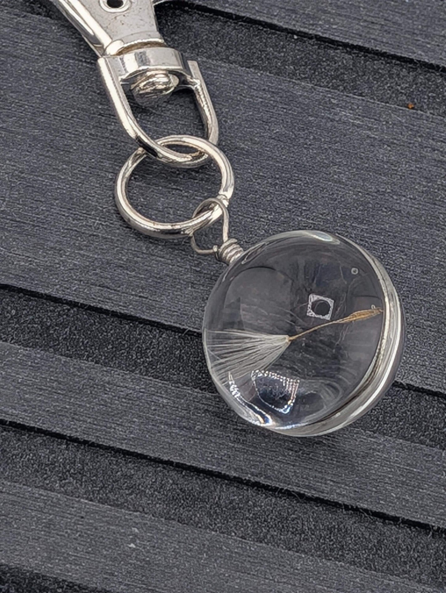 A clear glass keychain with a metal loop and a small white flower-like object attached to it, resting on a dark surface.