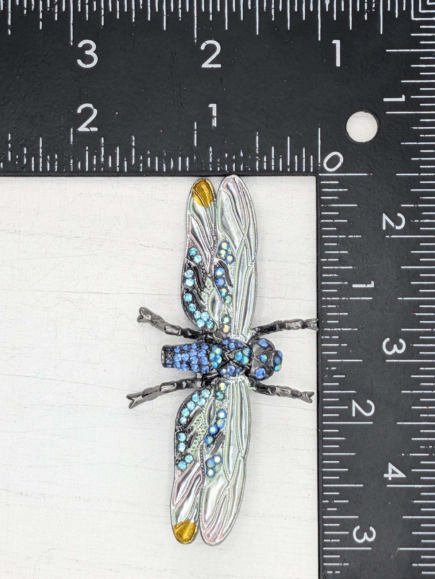 A close-up image of a decorative dragonfly brooch with blue and silver accents, placed on a ruler for size comparison.