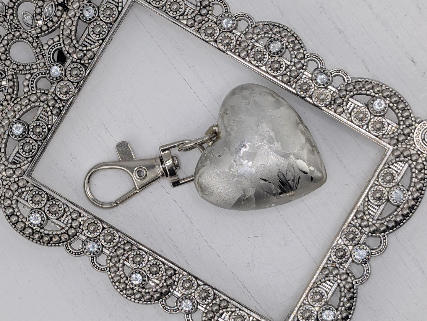 A silver heart-shaped charm with a silver clasp is displayed in a decorative silver frame with intricate designs.