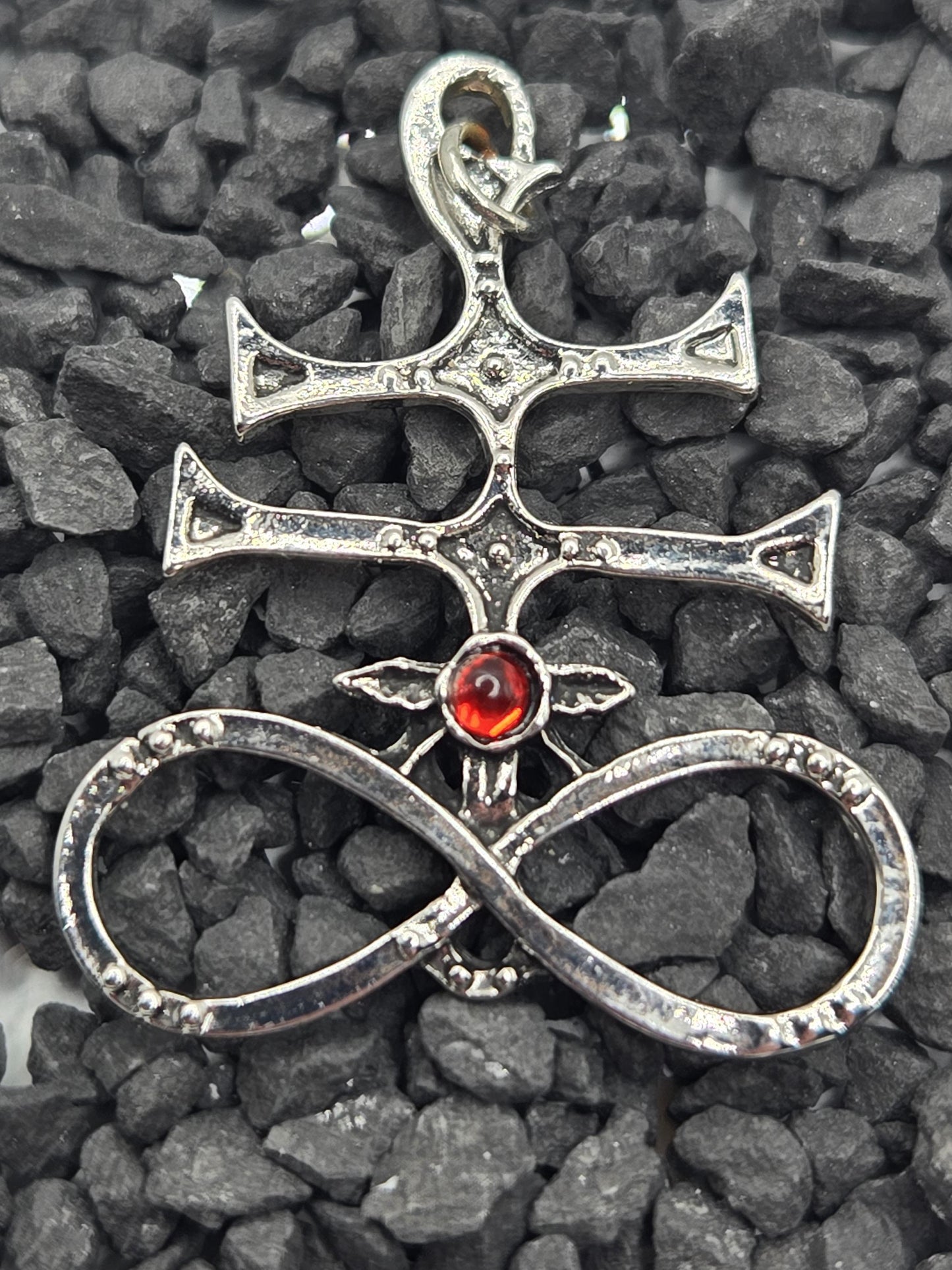 A silver pendant with a red gemstone, featuring an intricate design, is resting on a bed of small rocks.