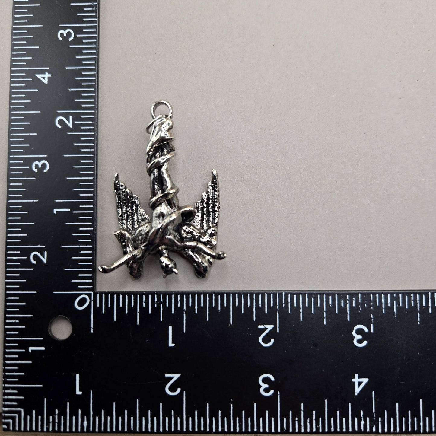 A silver-colored metal charm or pendant is placed next to a ruler on a gray surface.