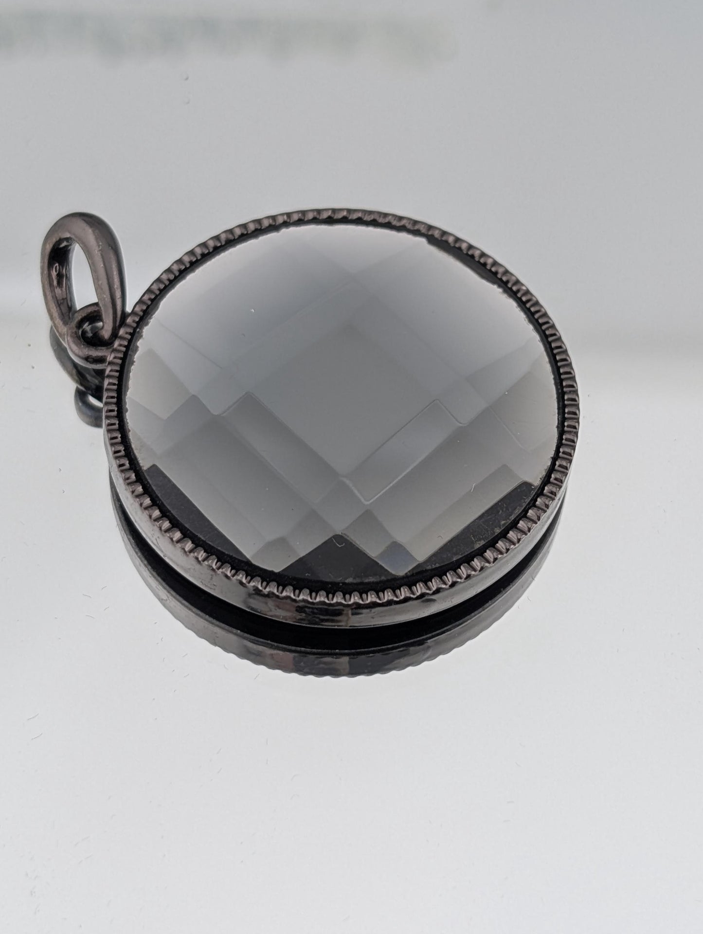 A round pendant with a black metal frame and a clear glass center, resting on a white surface.