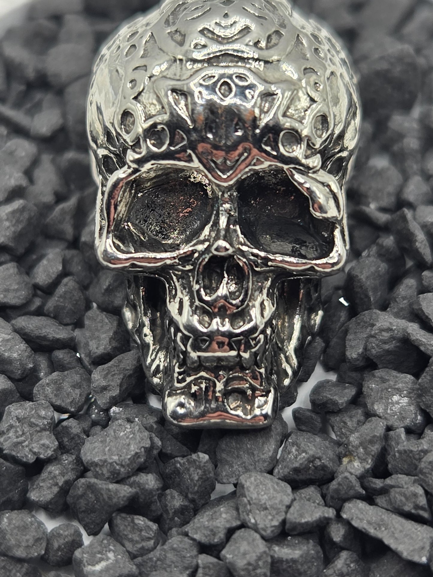A close-up image of a silver-colored metal skull lying on a bed of small gray rocks.