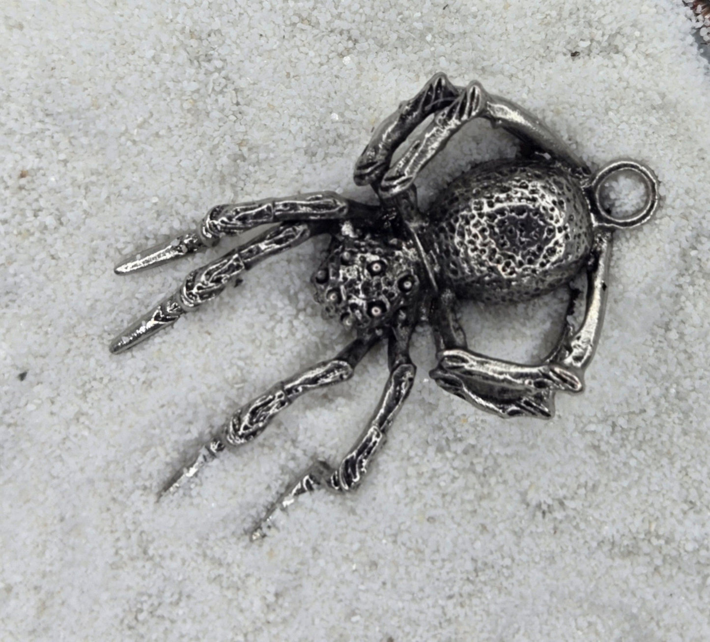 A detailed silver-colored metal spider pendant is lying on a snowy surface.