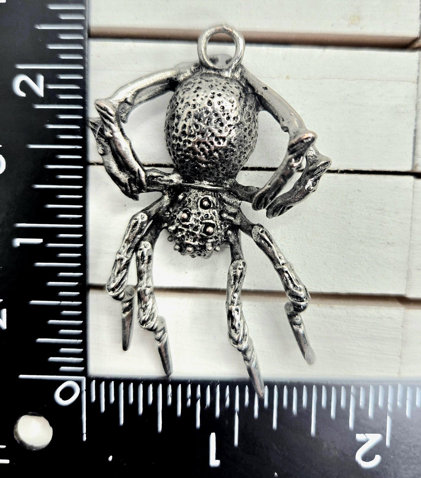 A close-up image of a detailed metal spider pendant, with a ruler for scale placed next to it.