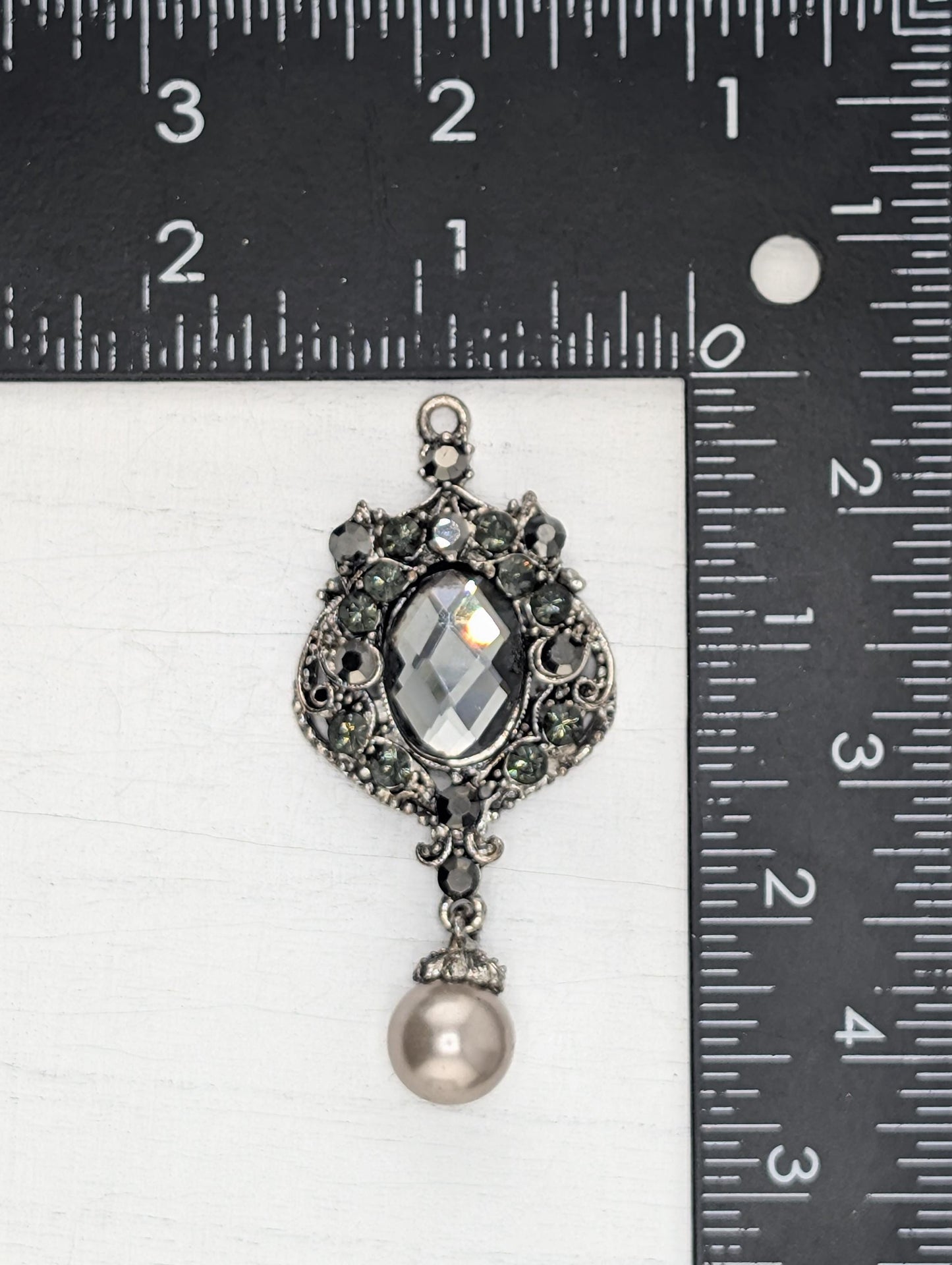 A silver-toned pendant with a crystal and pearl design, placed next to a ruler for size comparison.