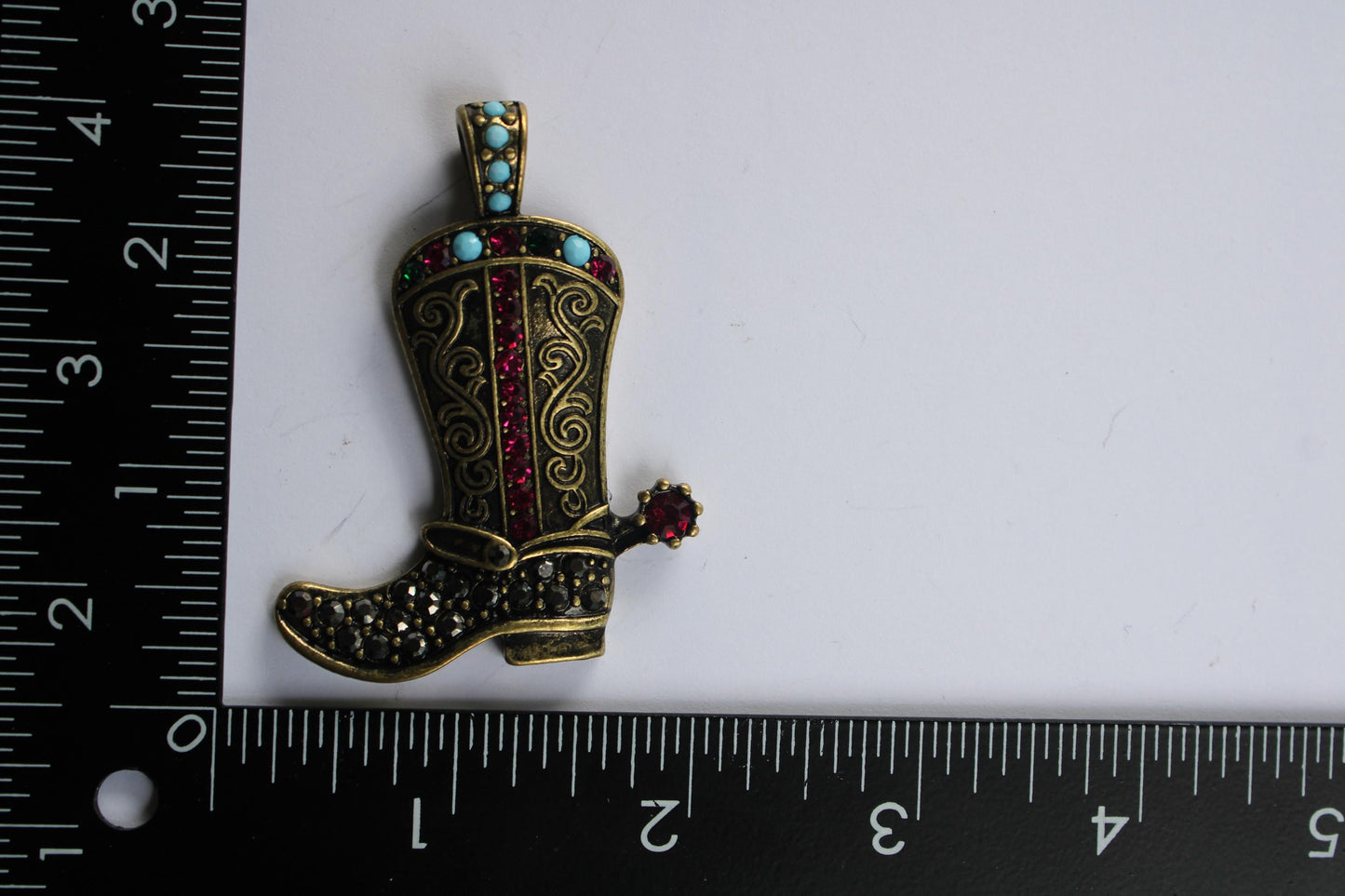 A decorative cowboy boot brooch with a red gemstone on the heel, placed next to a ruler for size reference.