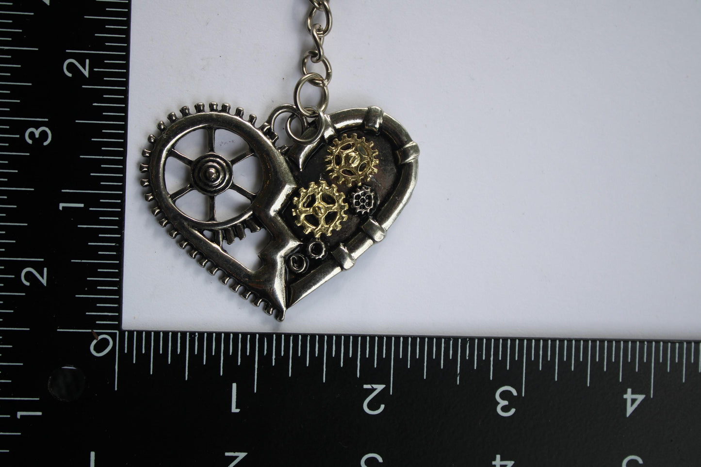 A keychain with a heart-shaped metal piece and gears attached to it, placed next to a ruler for size reference.