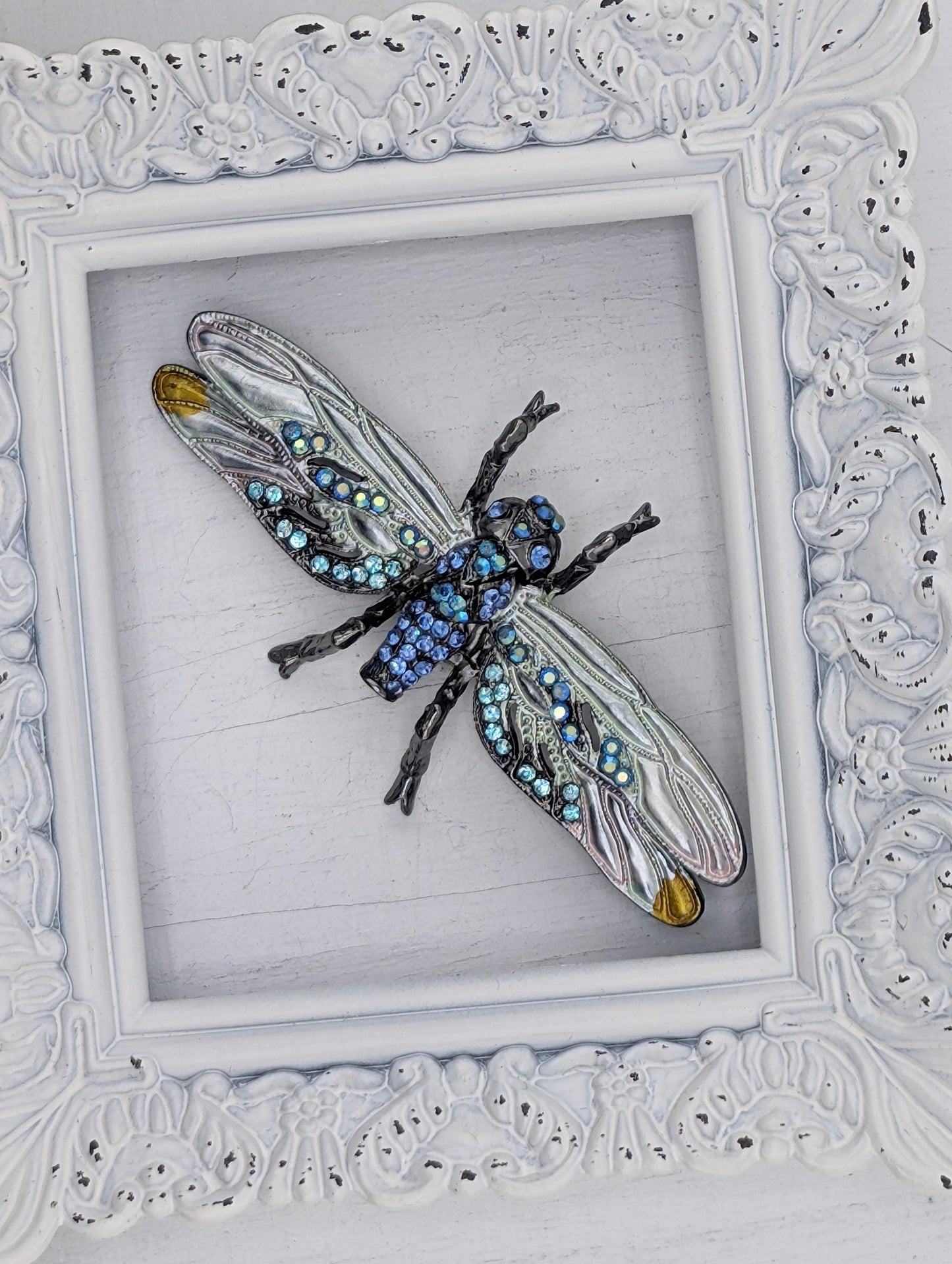 A decorative frame containing a large, intricately designed blue and silver insect, possibly a dragonfly or butterfly, with numerous small jewels or gemstones adorning its wings and body.