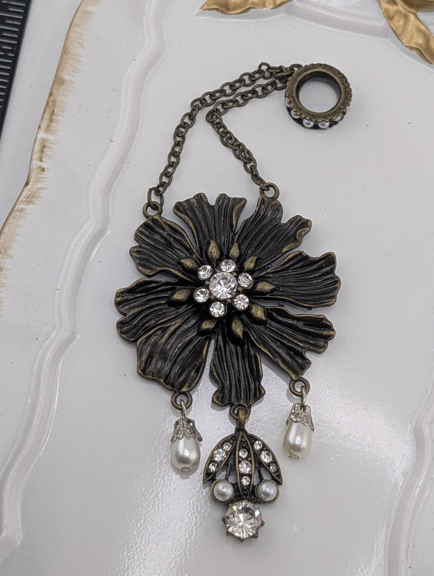 A decorative pendant with a large flower-shaped design, adorned with pearls and crystals, is displayed on a white surface.