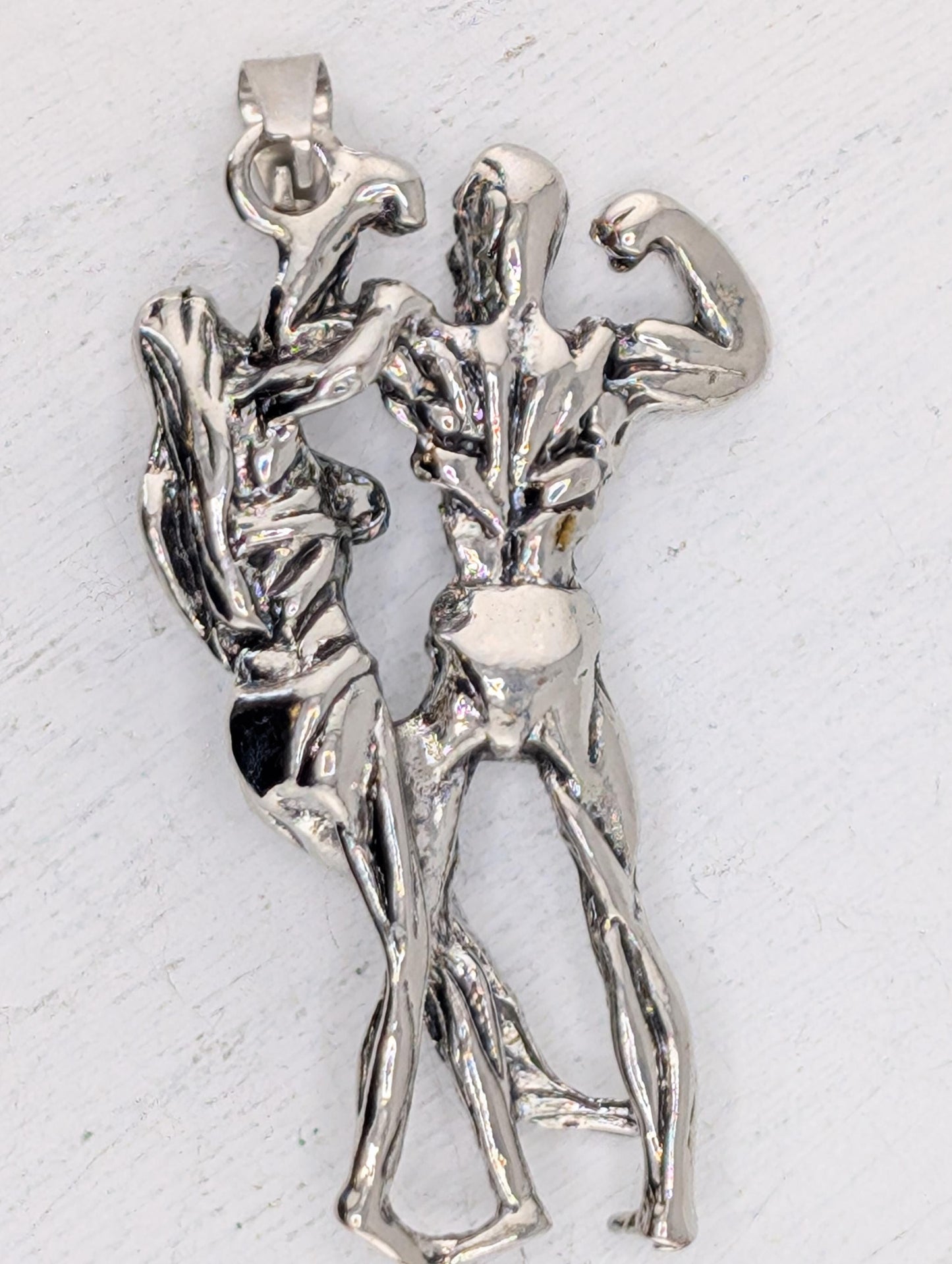 A silver-colored pendant in the shape of a muscular man and woman, with the man appearing to be flexing his muscles.