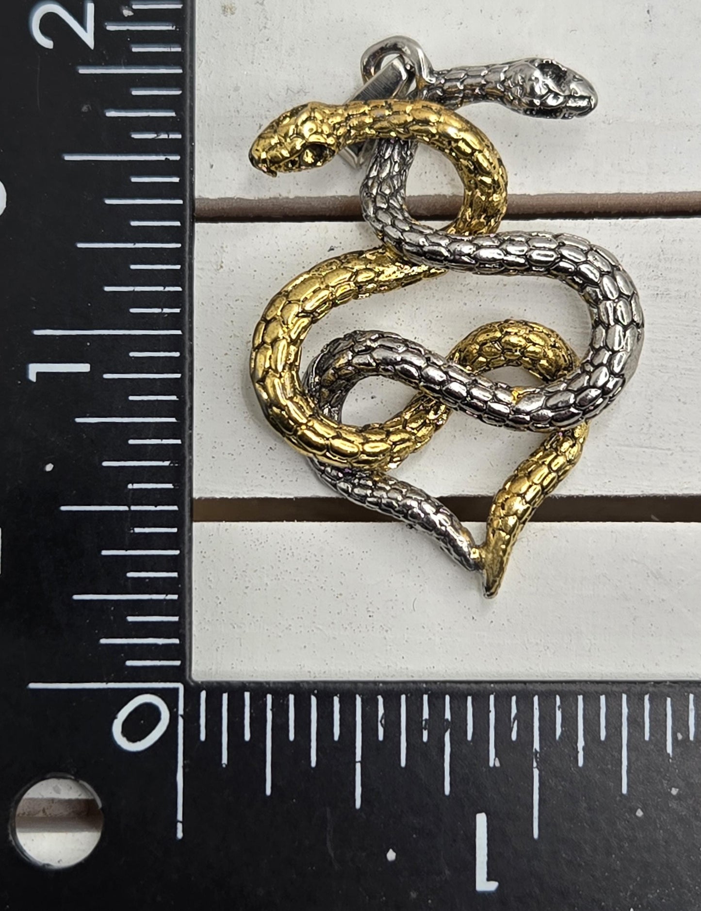 A close-up image of a pendant in the shape of a snake, with a gold and silver color scheme. The pendant is placed next to a ruler for size reference.