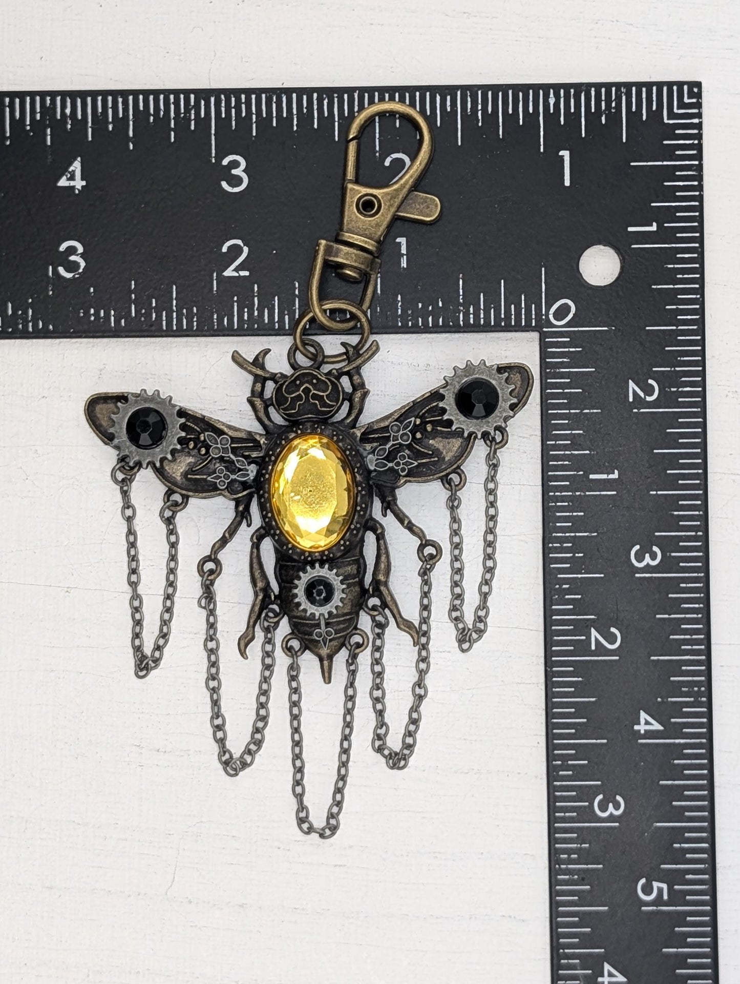 A detailed metal keychain in the shape of a bee, with a yellow gemstone in the center and chains hanging from its wings. The keychain is placed next to a ruler for size comparison.