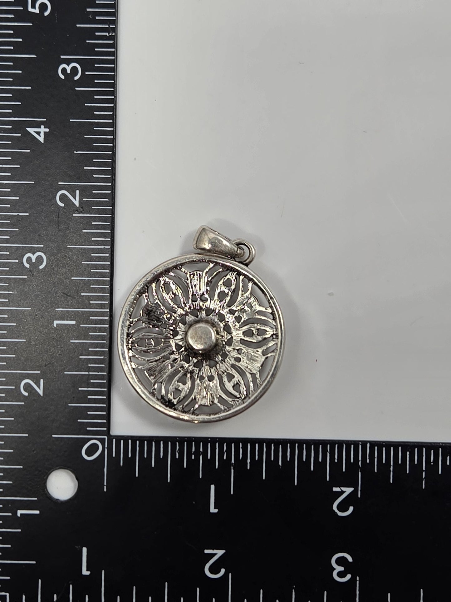 A silver-colored pendant with an intricate floral design is placed next to a ruler on a white surface.