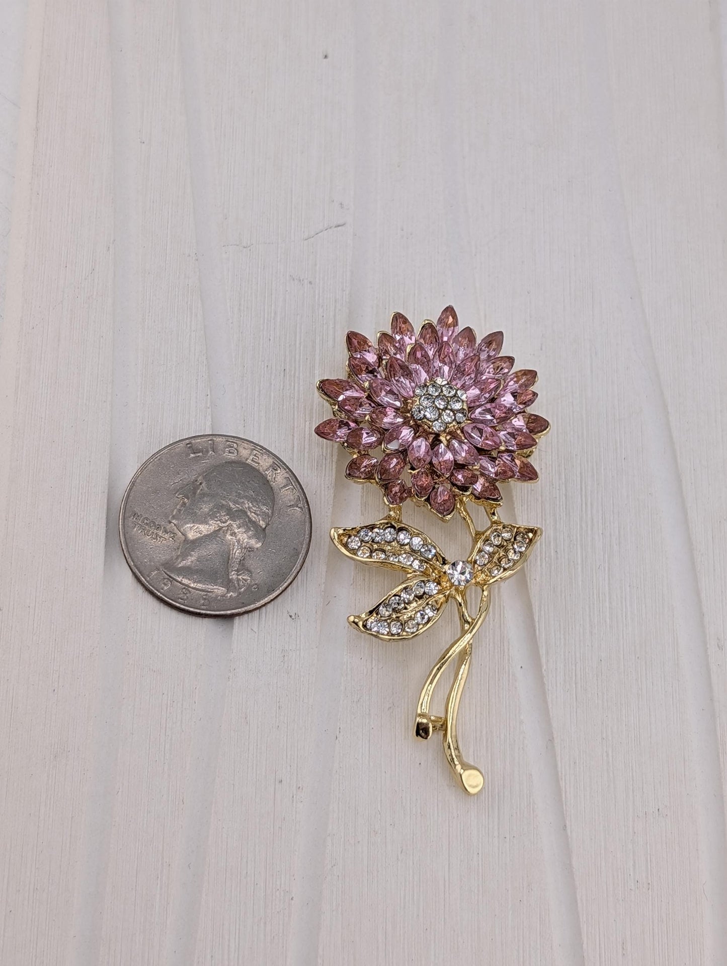 A pink flower brooch with sparkling gemstones is placed next to a US quarter on a white wooden surface.