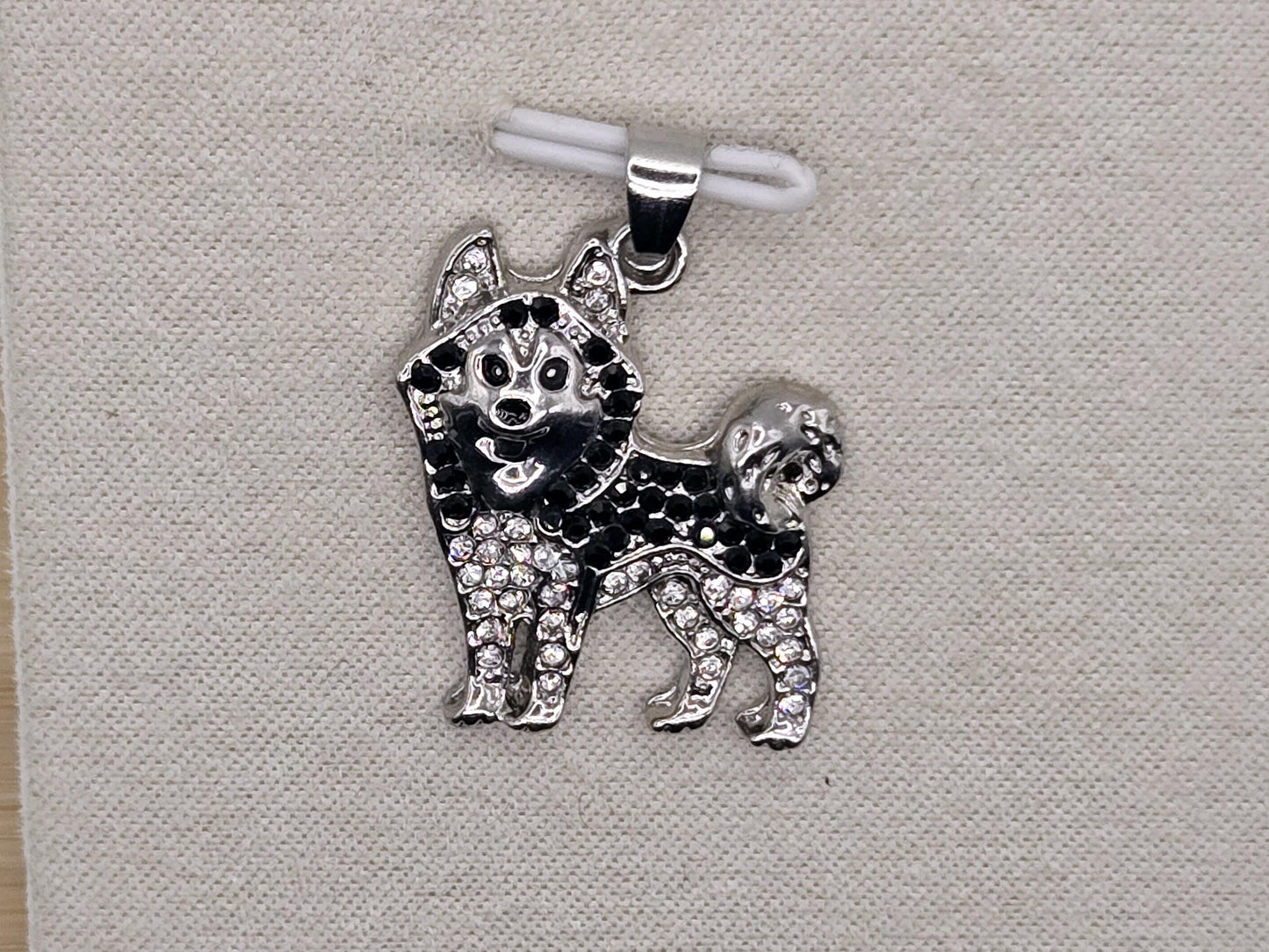 A silver-colored charm in the shape of a dog, adorned with small diamonds or crystals, is displayed on a white fabric background.