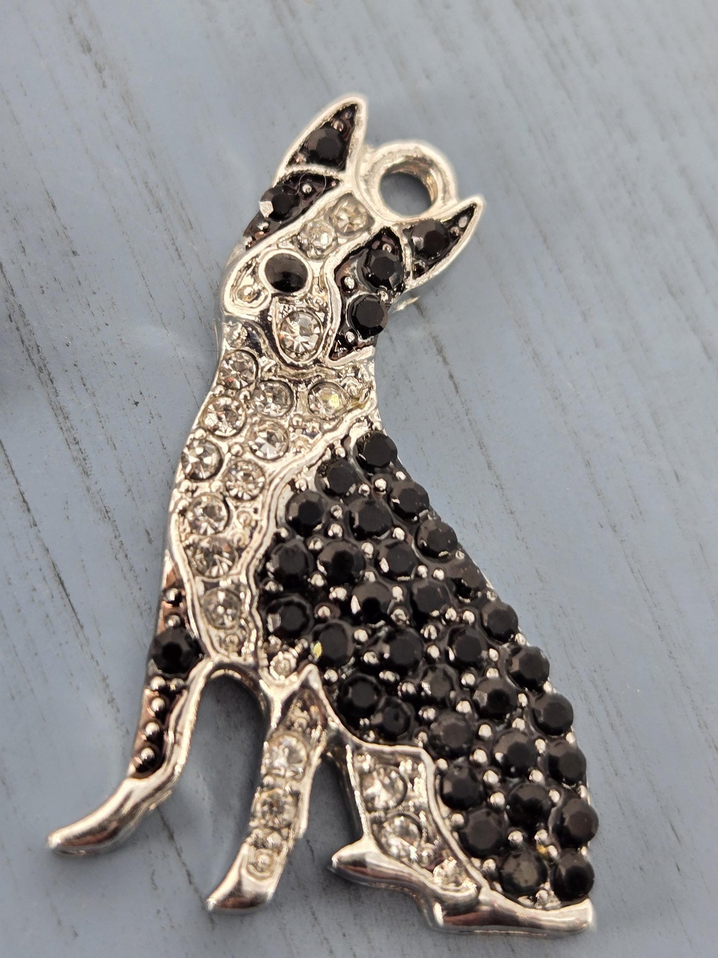 A silver-colored pendant or charm in the shape of a dog, adorned with black rhinestones or crystals.