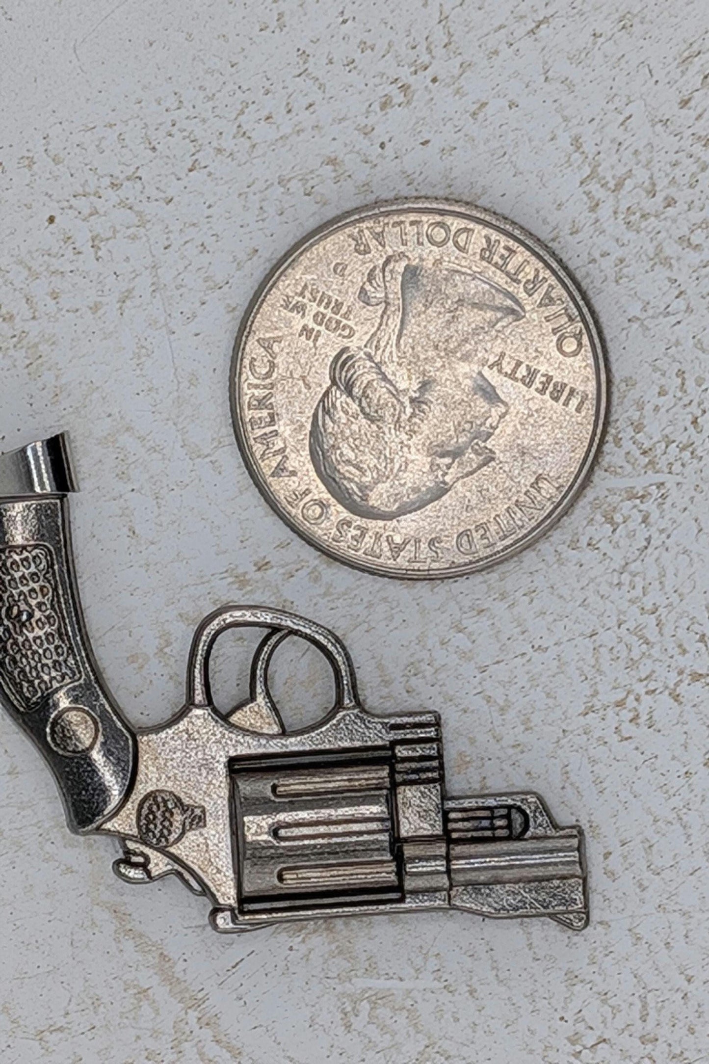 A silver-colored toy gun is placed next to a United States quarter dollar coin on a white surface.