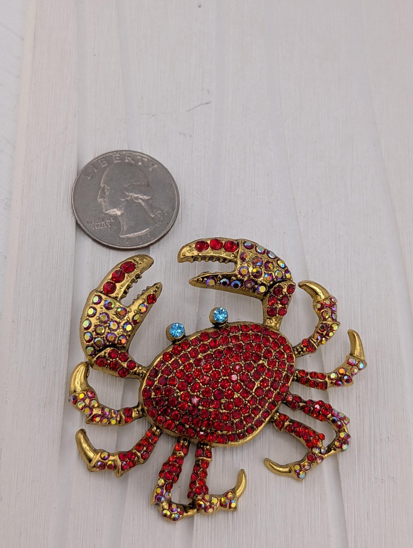 A shiny, ornate crab brooch with red and gold jewels is placed next to a United States quarter coin on a white surface.