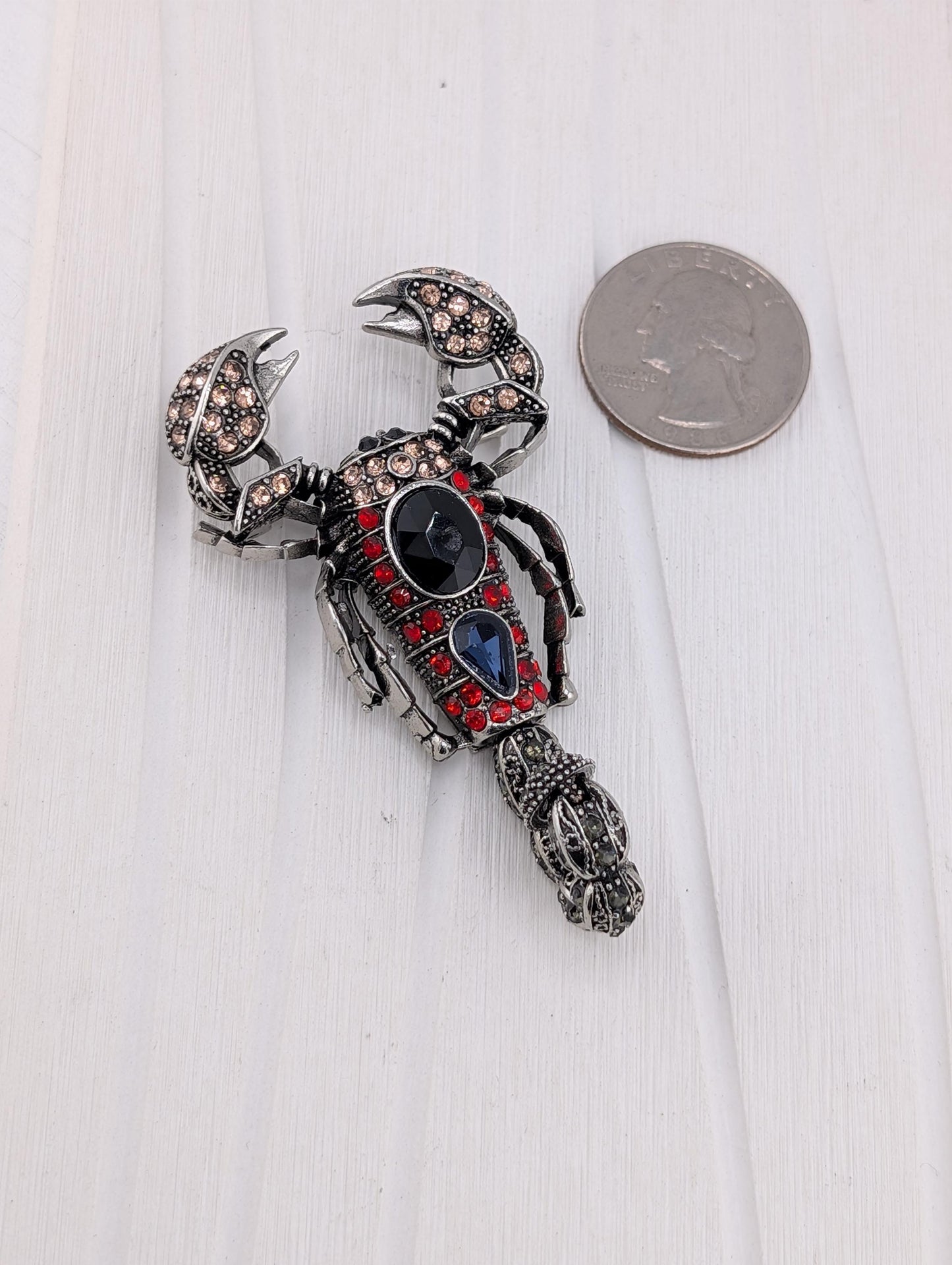 A decorative metal lobster brooch with a red and black color scheme, adorned with gemstones, is placed next to a US quarter on a white surface.