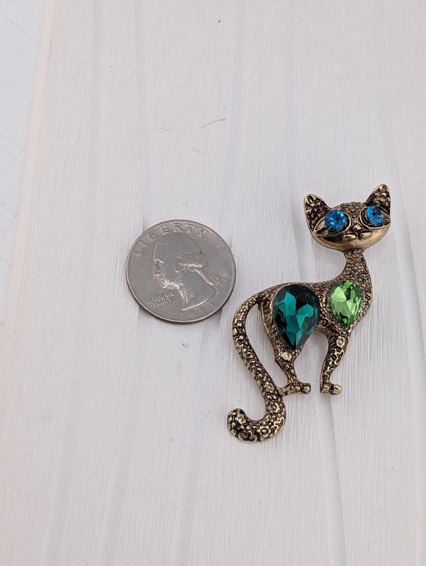 A shiny, ornate cat-shaped brooch made of metal and adorned with colorful gemstones is placed next to a US quarter on a white surface.