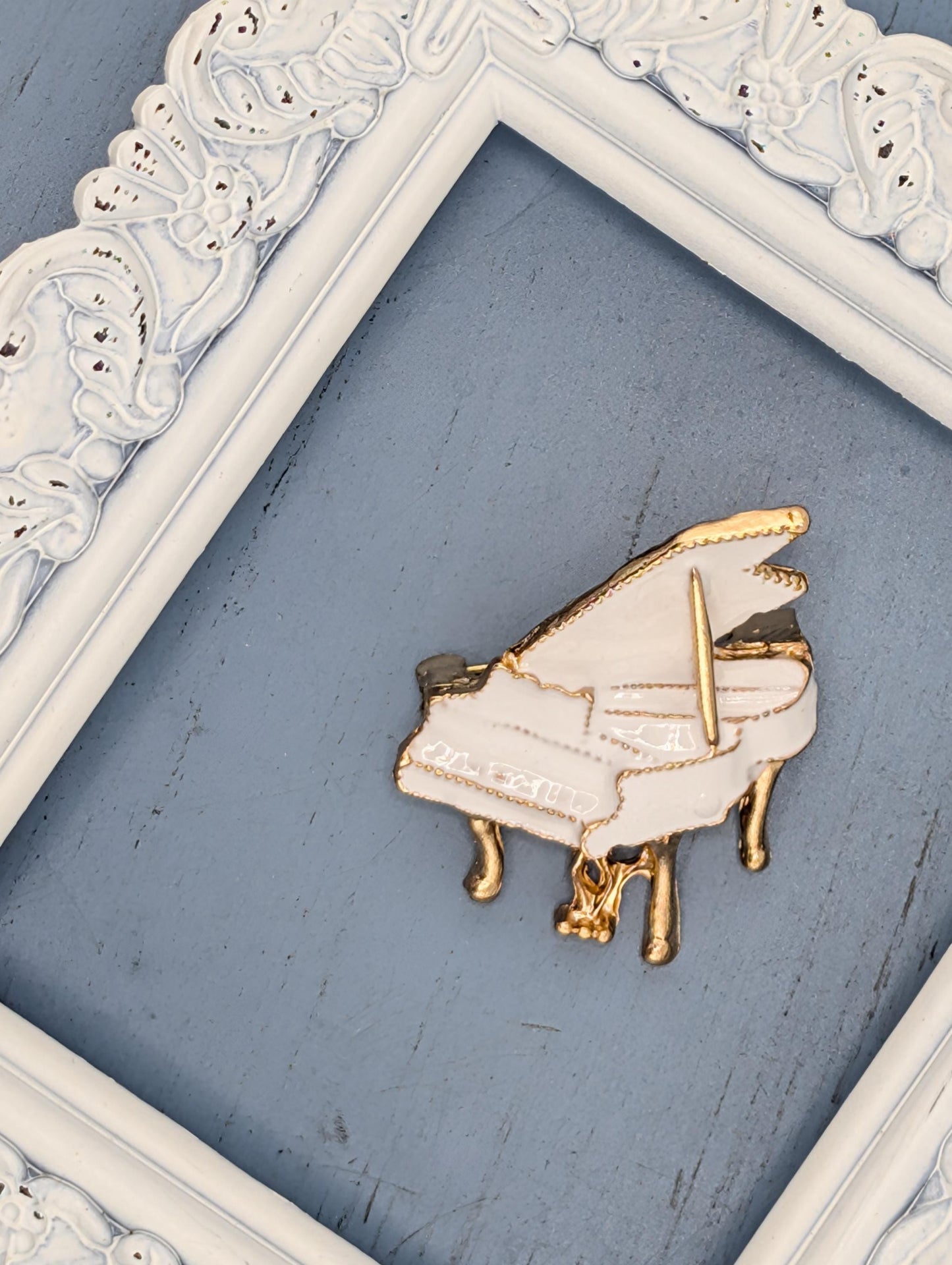 A white and gold piano brooch is displayed on a blue surface, framed by a white ornate frame.