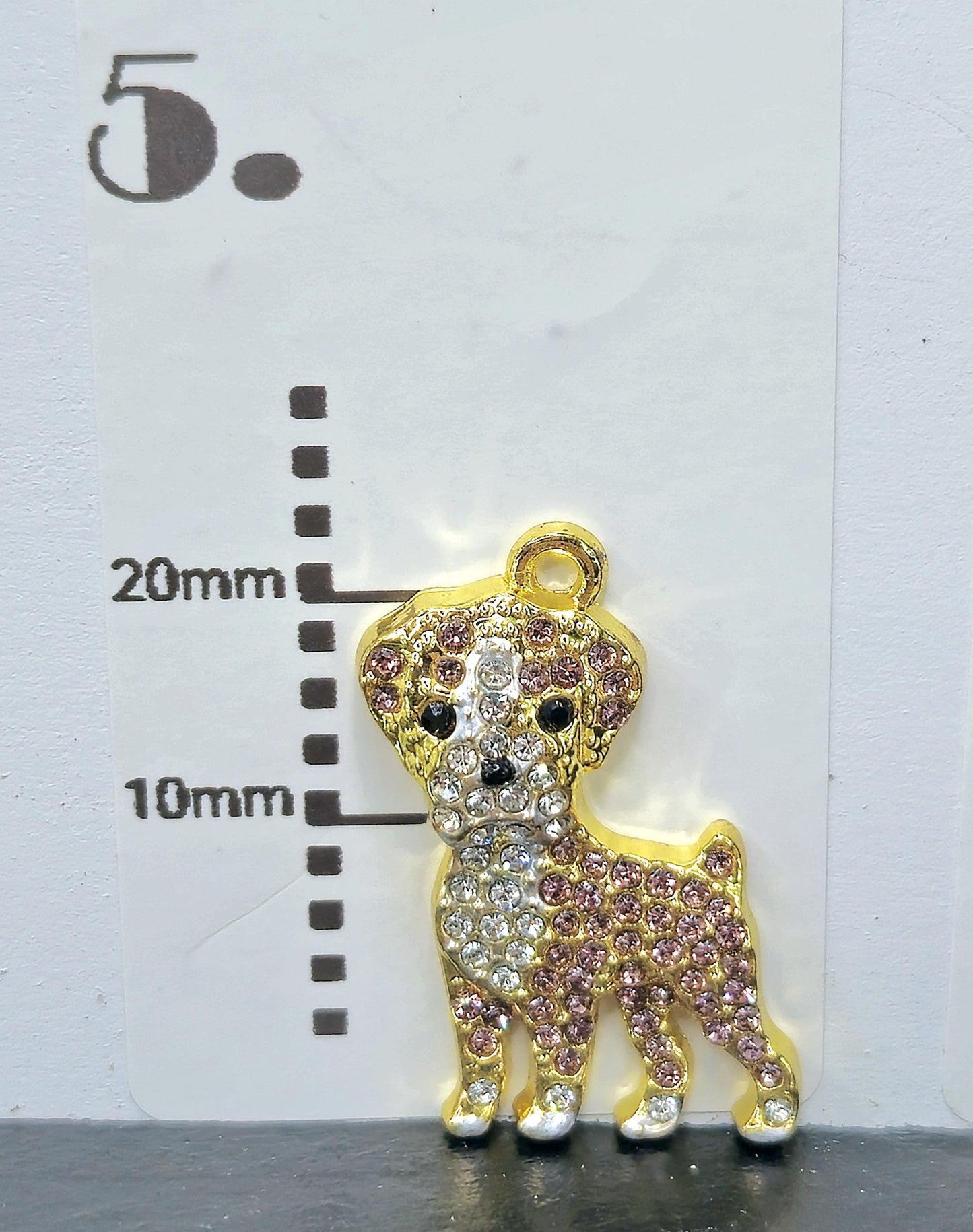 A gold-colored charm in the shape of a dog, adorned with rhinestones and a small bell, is placed next to a ruler for size comparison.