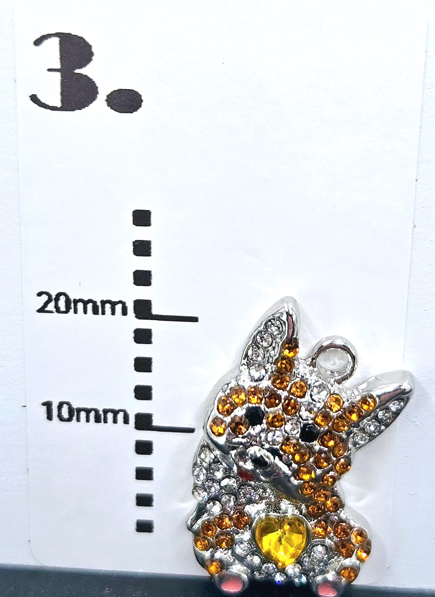 A pug charm with a yellow heart and sparkling rhinestones is shown next to a ruler indicating its size.