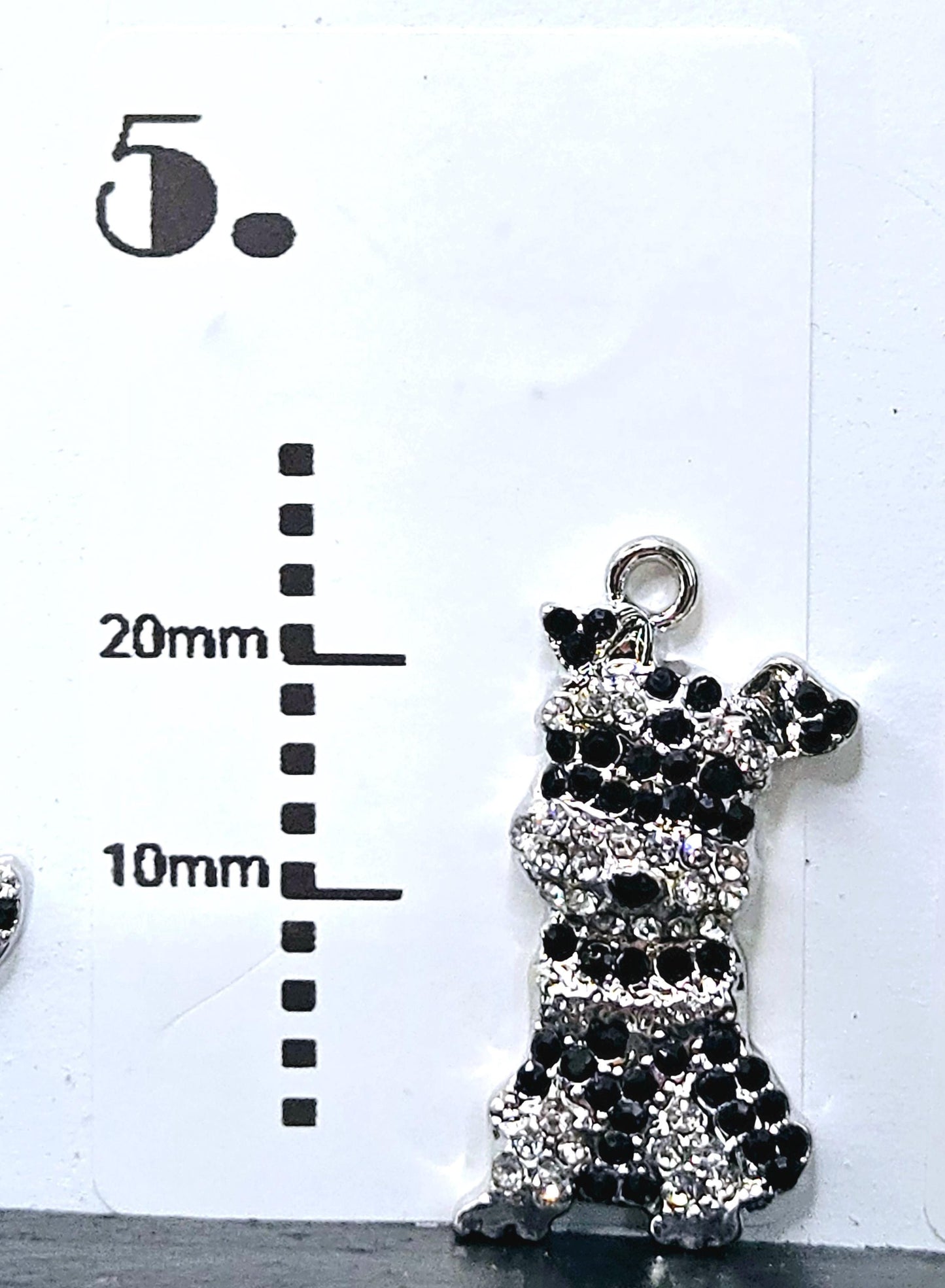 A close-up image of a pendant with a black and white design, featuring a small charm attached to it. The pendant appears to be made of metal and is placed next to a scale for size comparison.