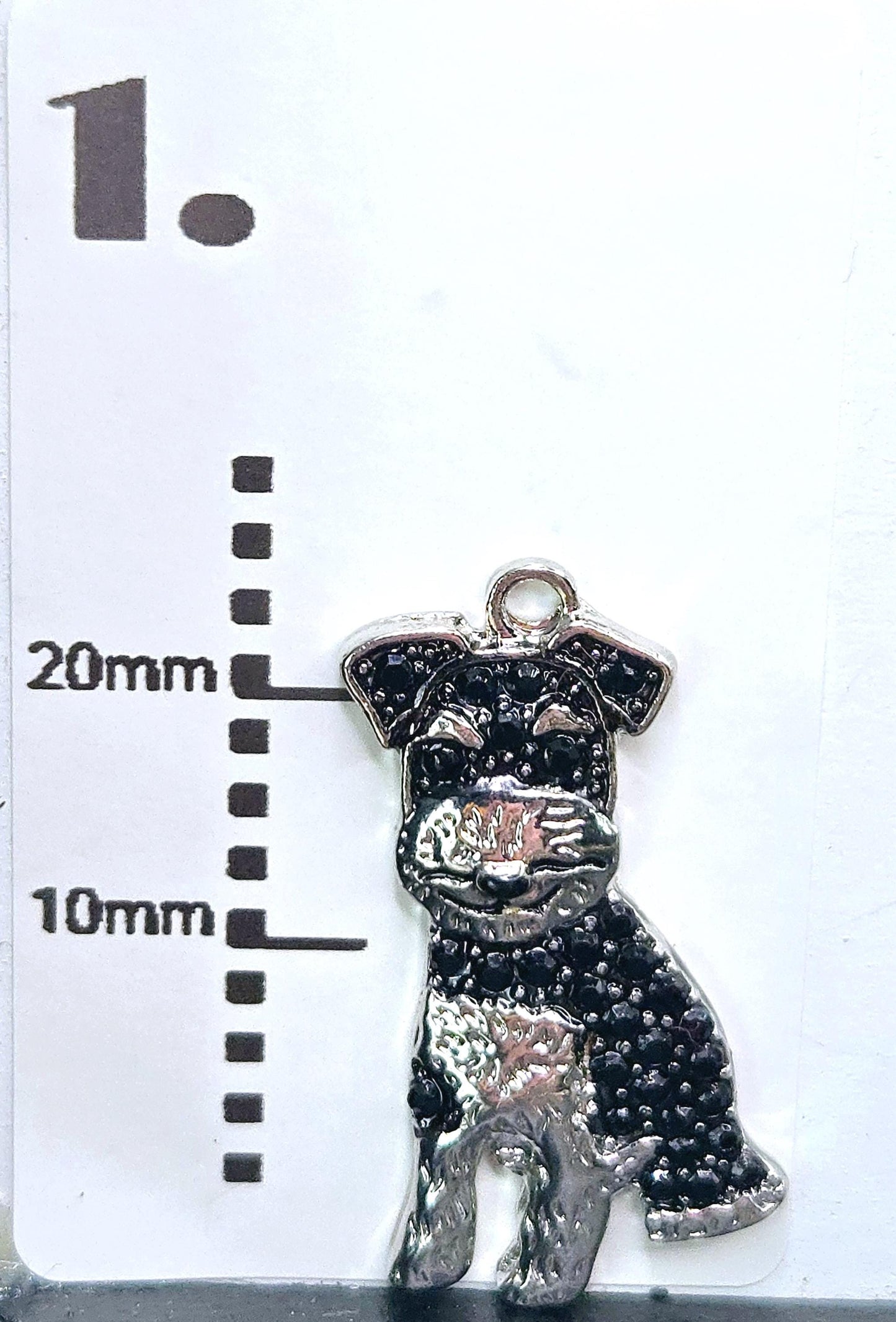 A silver charm in the shape of a dog, with a black and white pattern, is shown next to a ruler indicating its size.