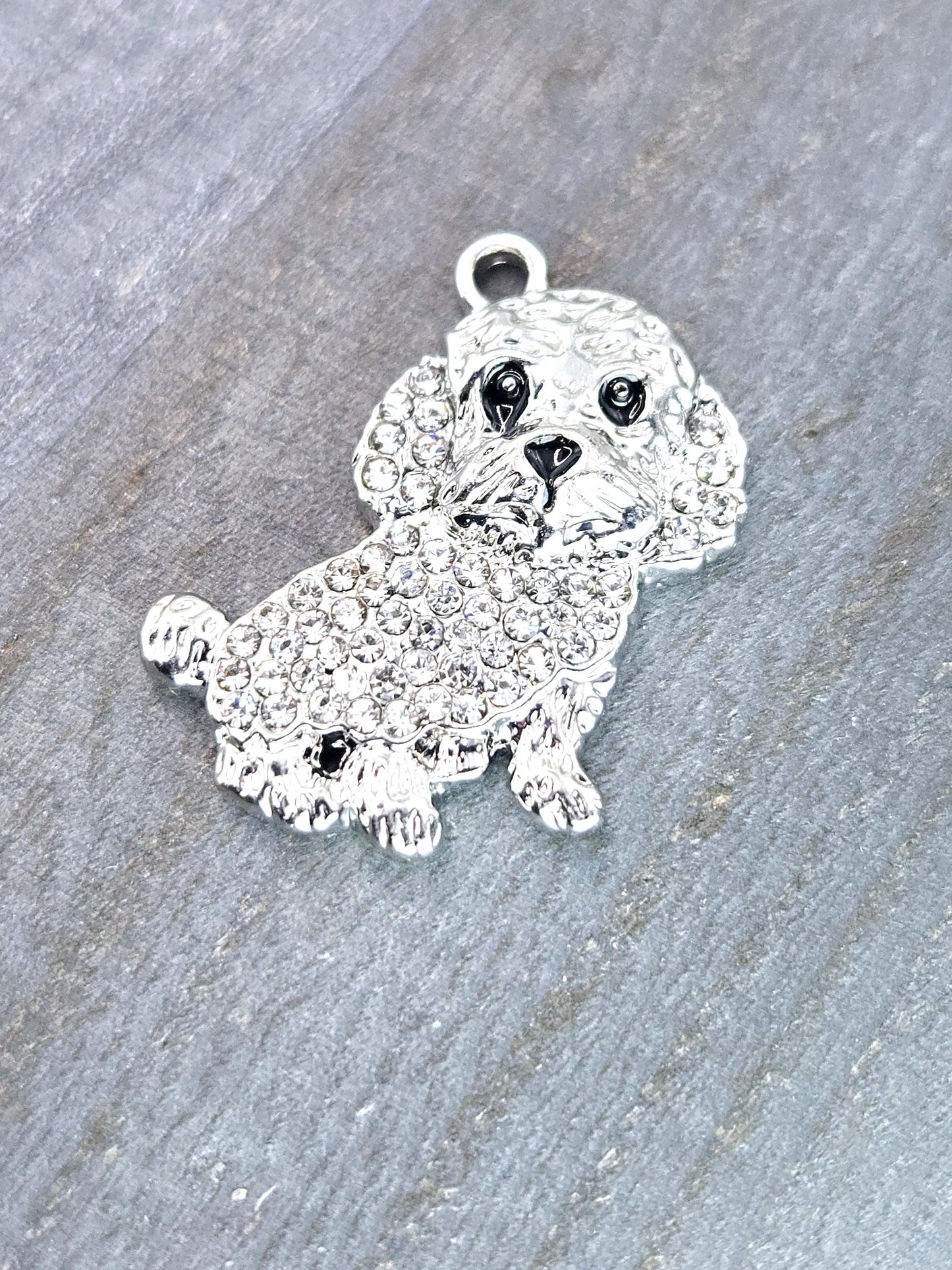A silver charm in the shape of a dog, with a sparkling crystal or rhinestone design, is resting on a textured gray surface.