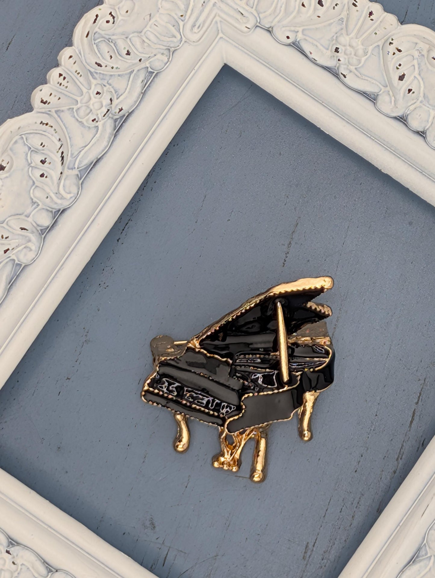 A decorative gold-colored piano brooch is displayed on a blue surface, framed by a white ornate frame.