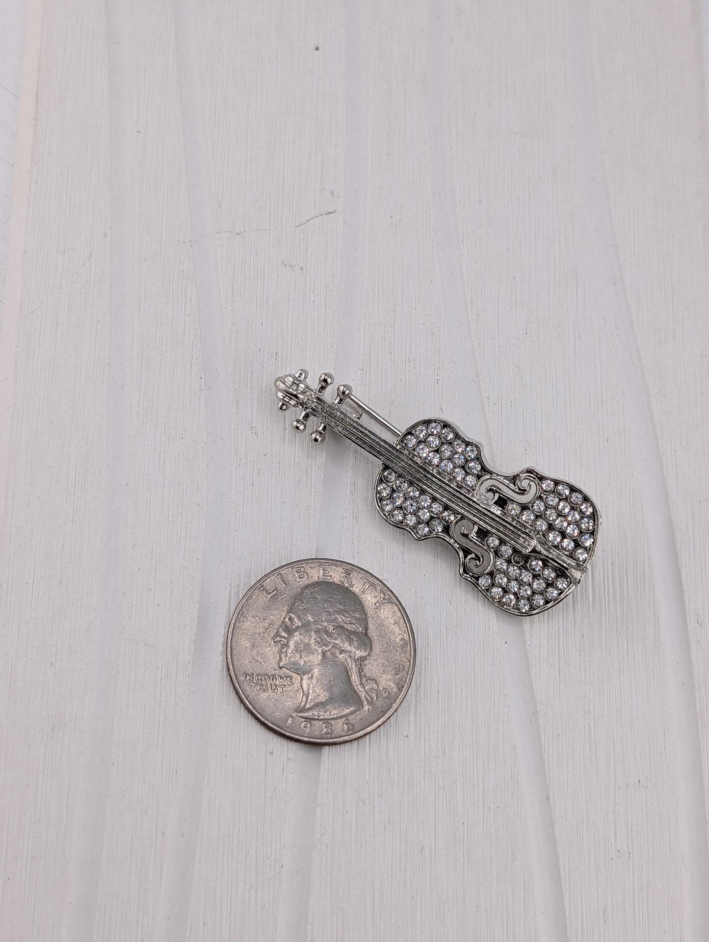 A silver-colored violin brooch with rhinestones is placed next to a United States quarter on a white wooden surface.