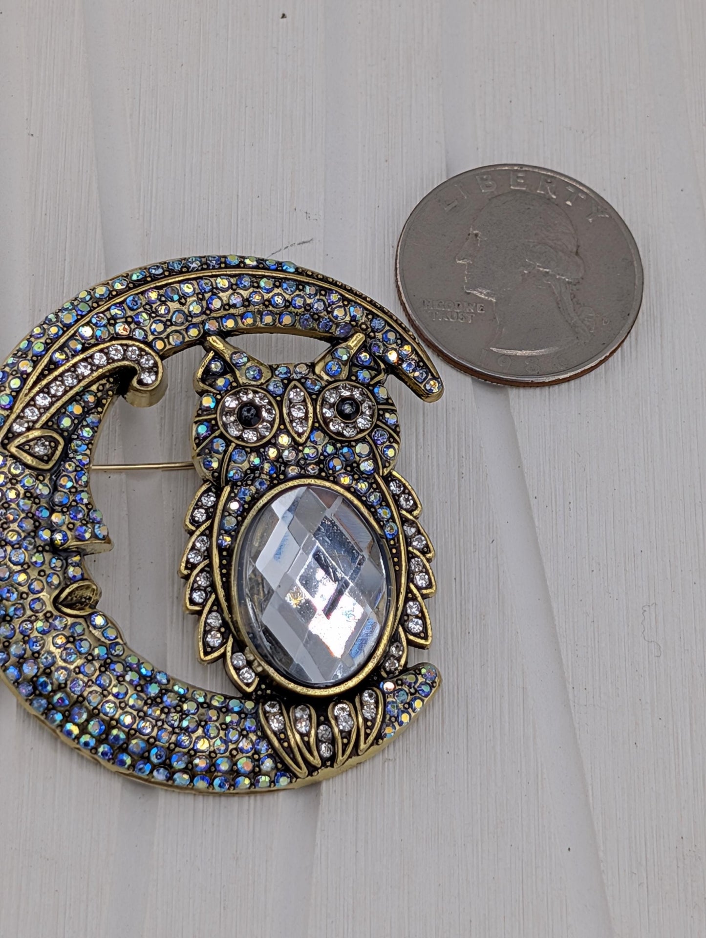 A brooch in the shape of a crescent moon, adorned with a large crystal owl, is placed next to a United States quarter.