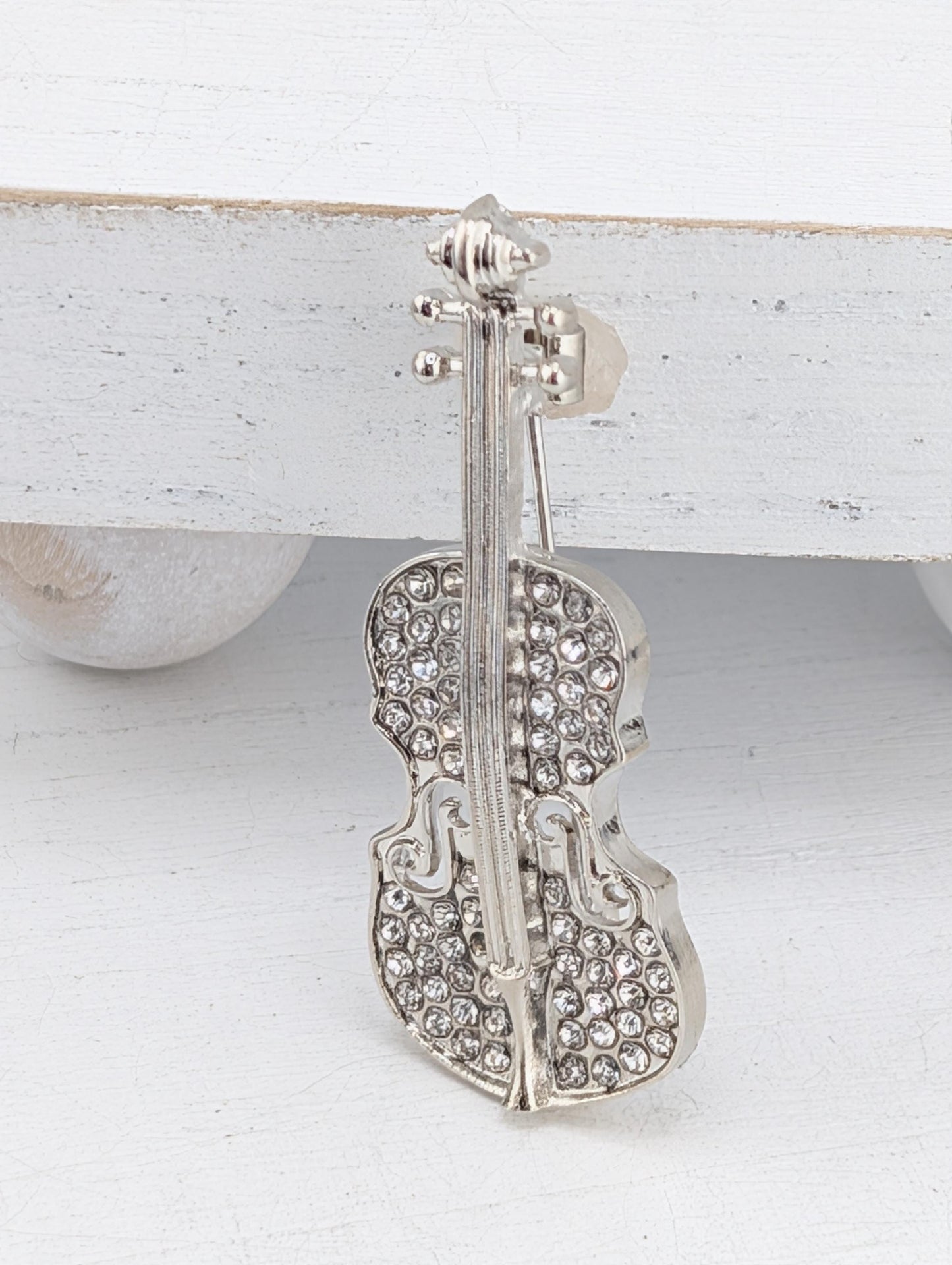 A silver-colored brooch or pin featuring a detailed design of a violin with rhinestones or crystals embedded in it.