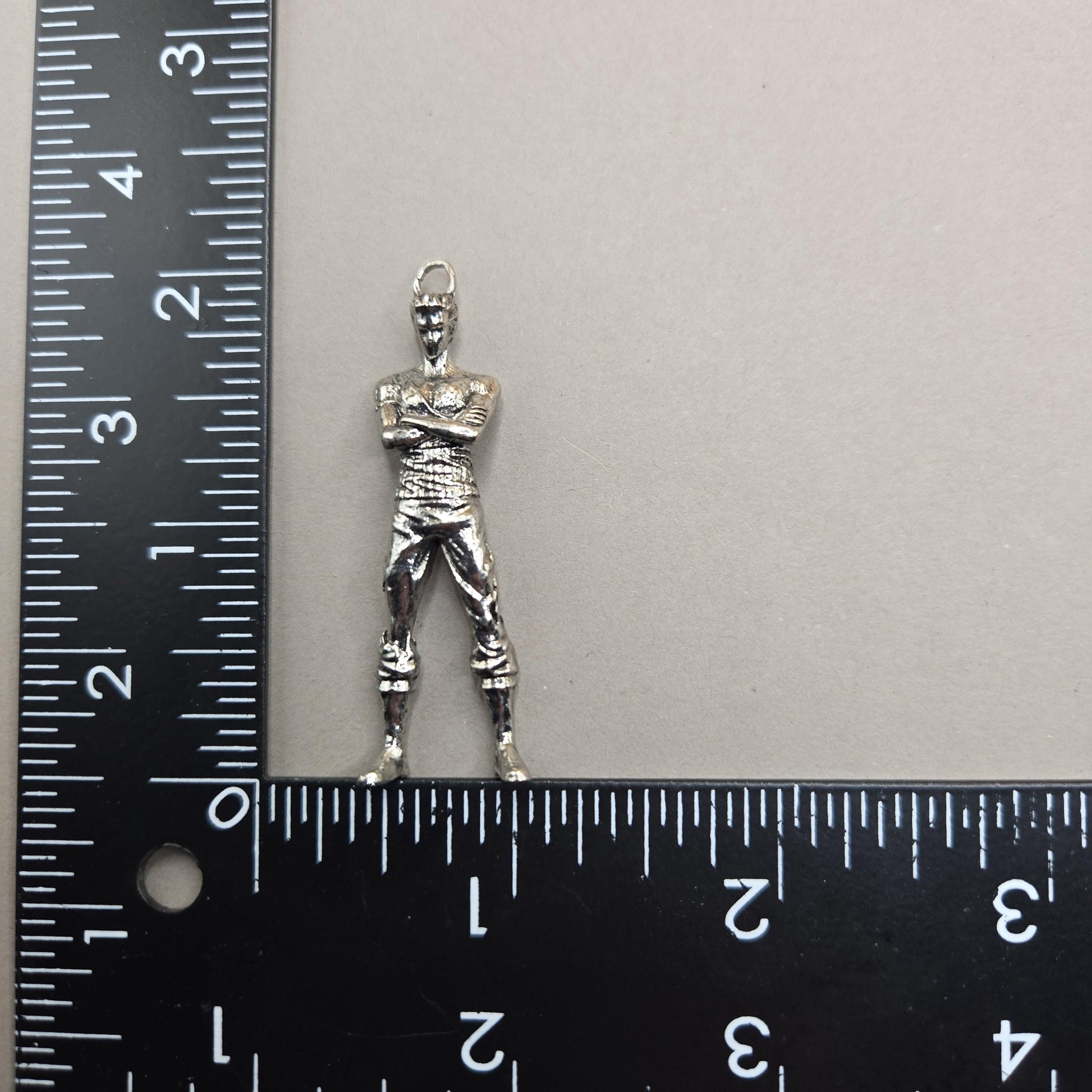 A silver-colored metal charm or pendant is placed next to a ruler on a gray surface.