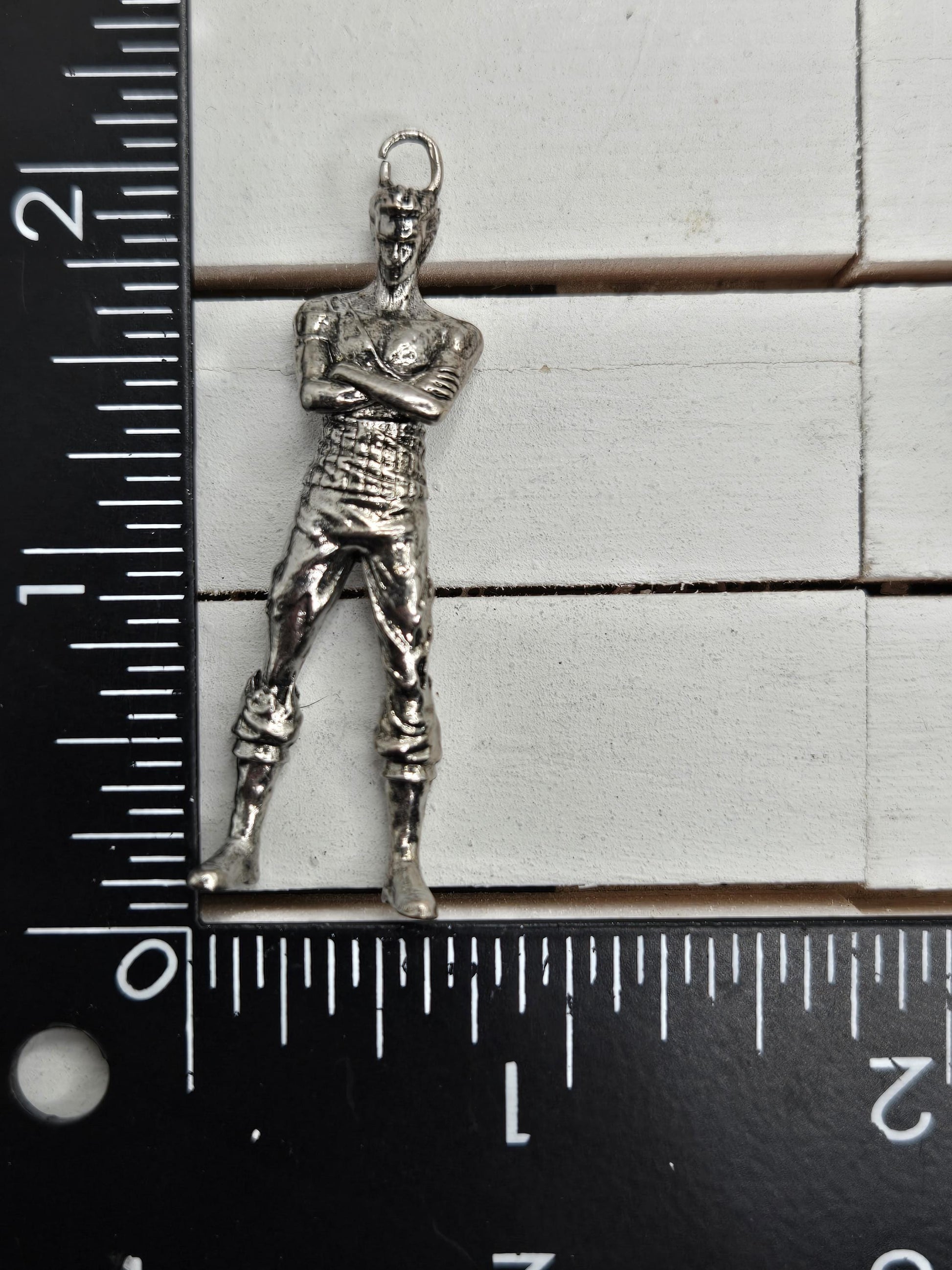 A silver-colored metal charm or pendant is placed next to a ruler on a white surface.