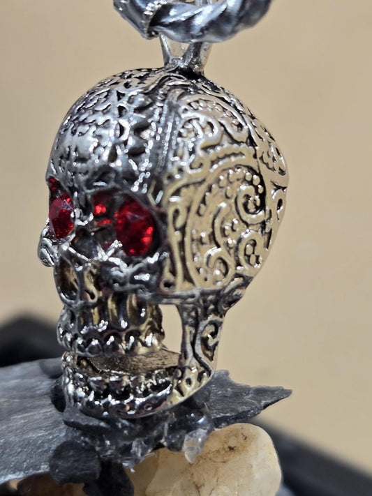 A close-up view of a silver-colored metal pendant with a red gemstone, resembling a skull or skull-shaped design.