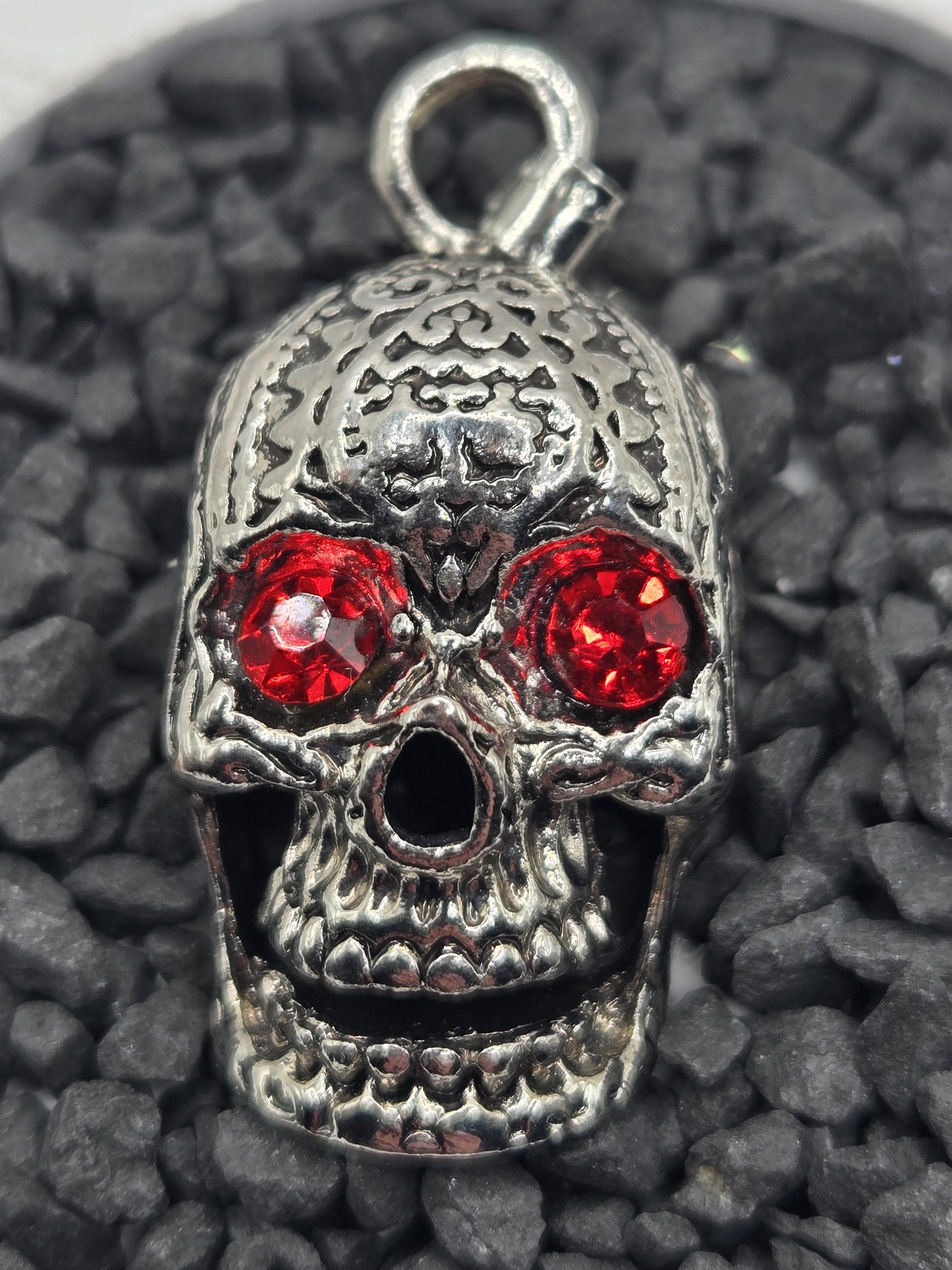 A silver skull pendant with red gemstones is shown against a dark background of small rocks.
