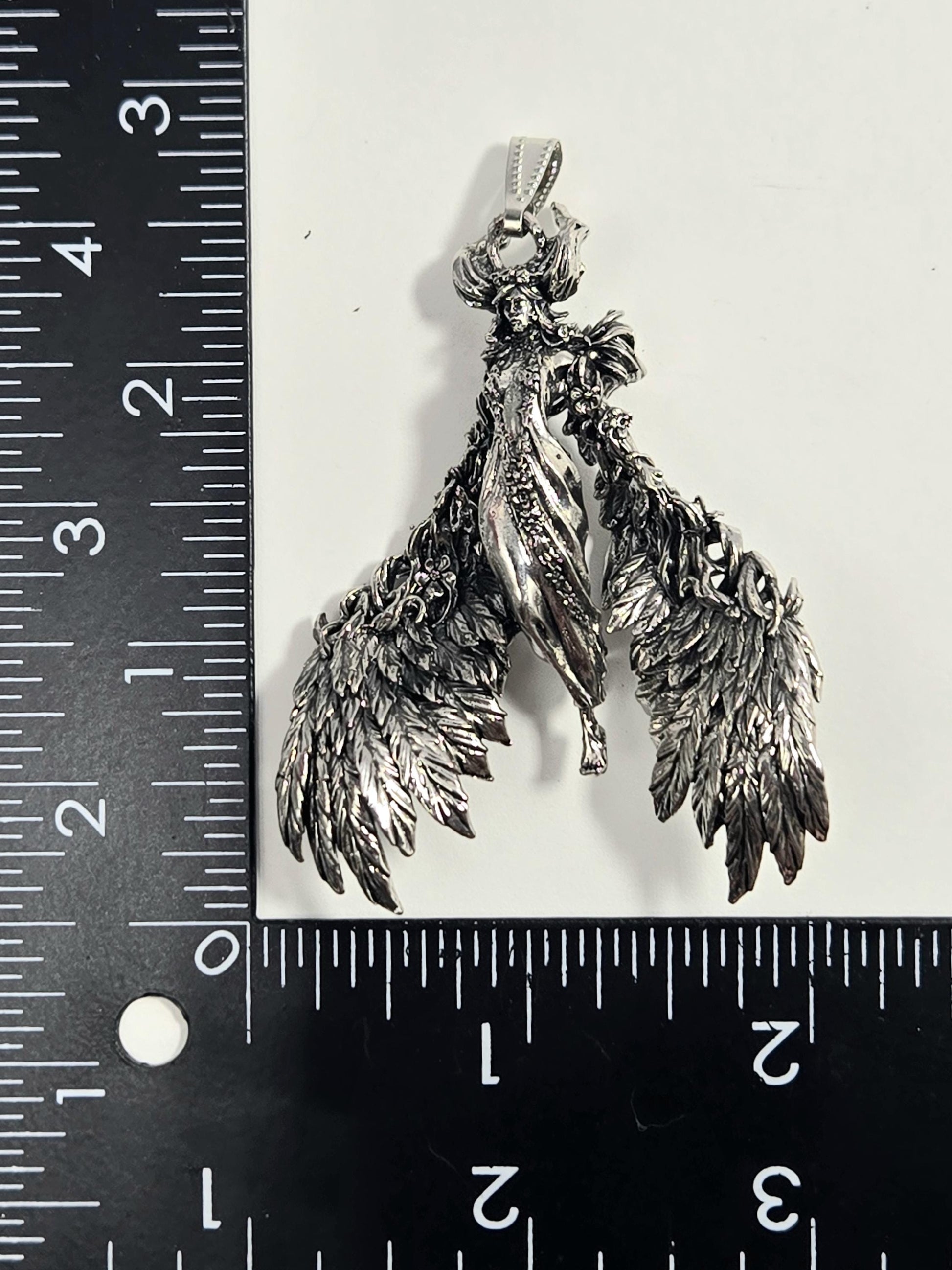 A silver-colored pendant in the shape of a winged figure, possibly a deity or angel, is being measured against a ruler.