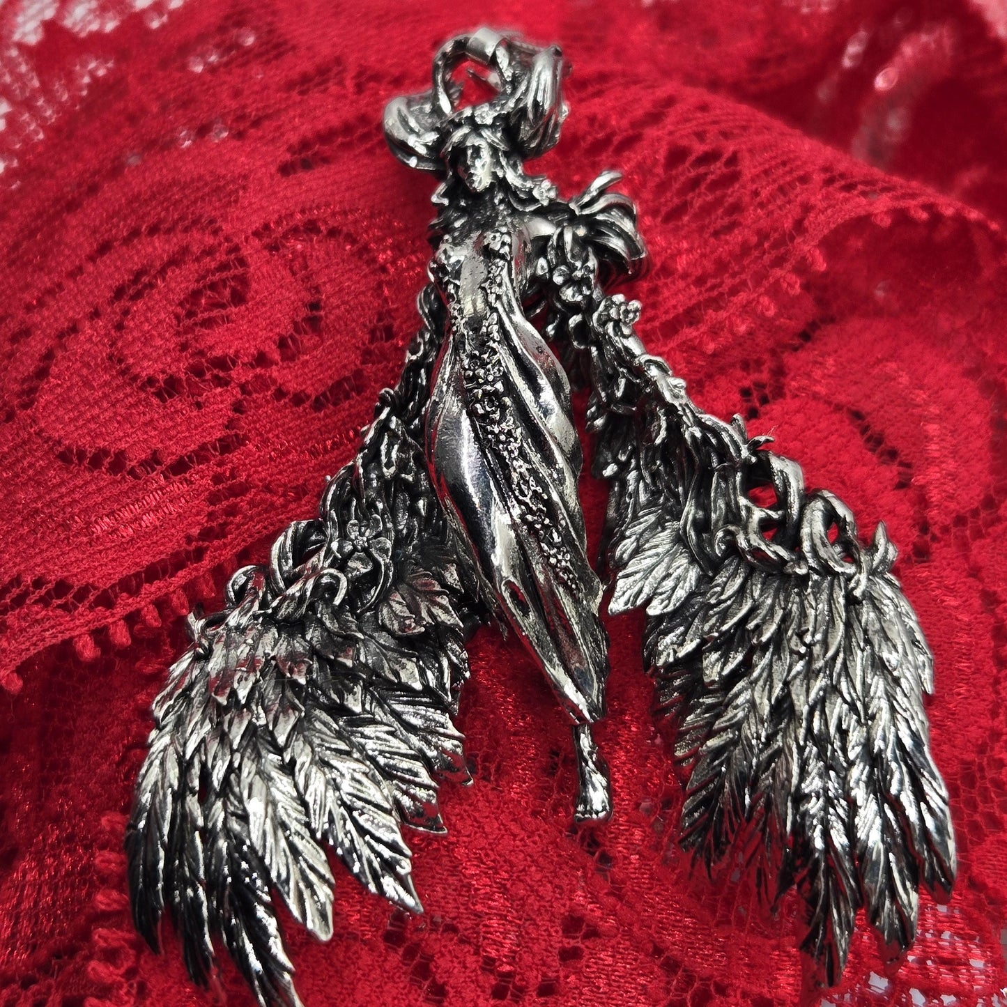 A close-up image of a silver, winged brooch or pendant resting on a red, lace-like fabric.