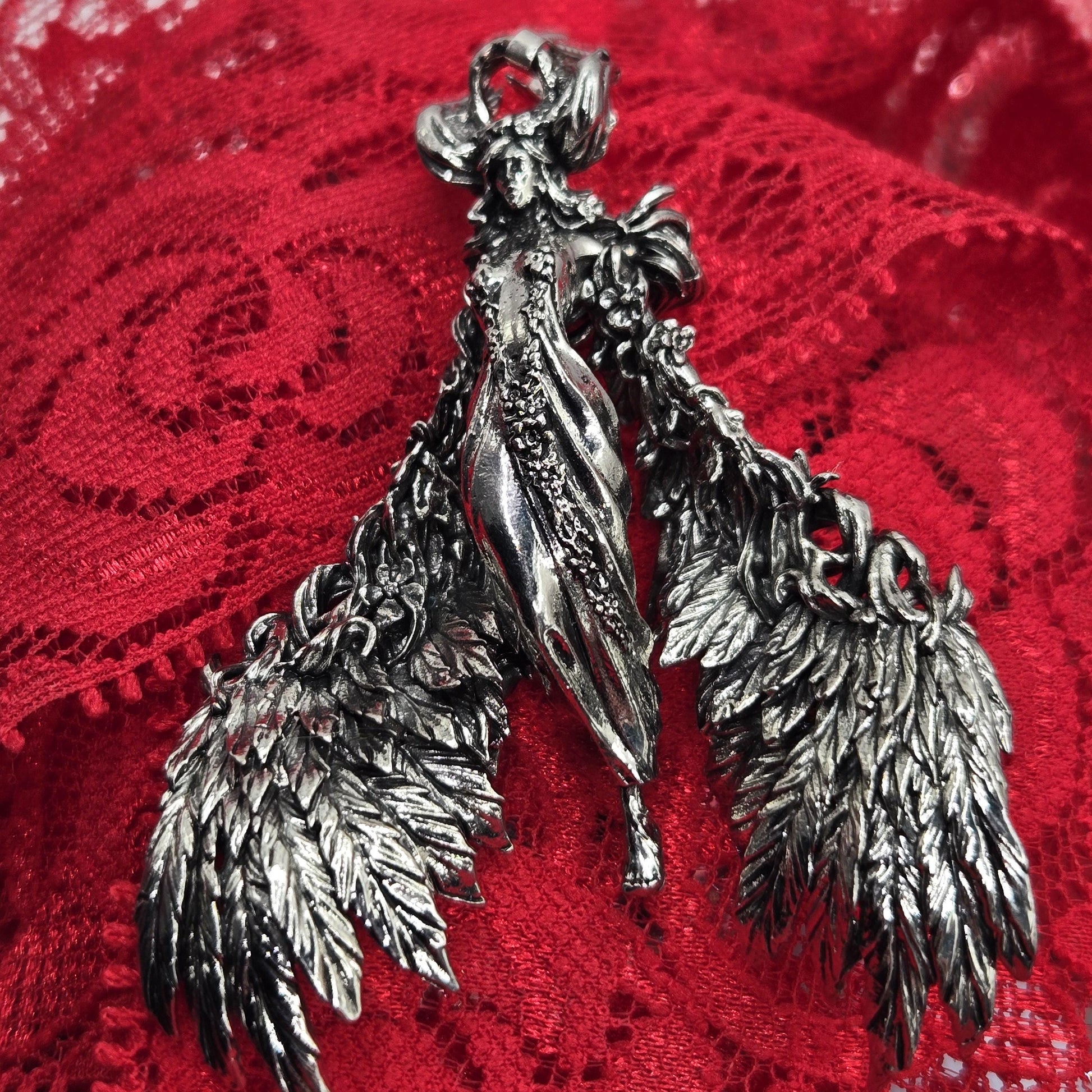 A close-up image of a silver, winged brooch or pendant resting on a red, lace-like fabric.