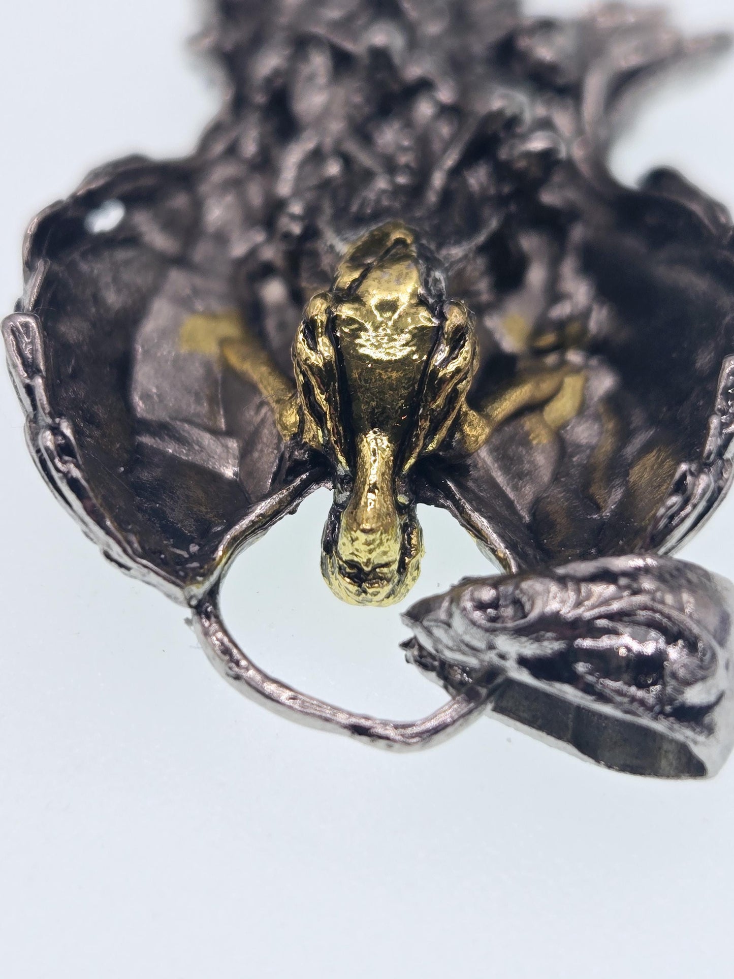 a close-up view of a unique, intricately designed metal piece, possibly a pendant or sculpture. The piece appears to be made of a combination of silver and gold materials, with a textured and ornate appearance. The image does not contain any text or other discernible objects, and the focus is solely on the detailed design of the metal piece.