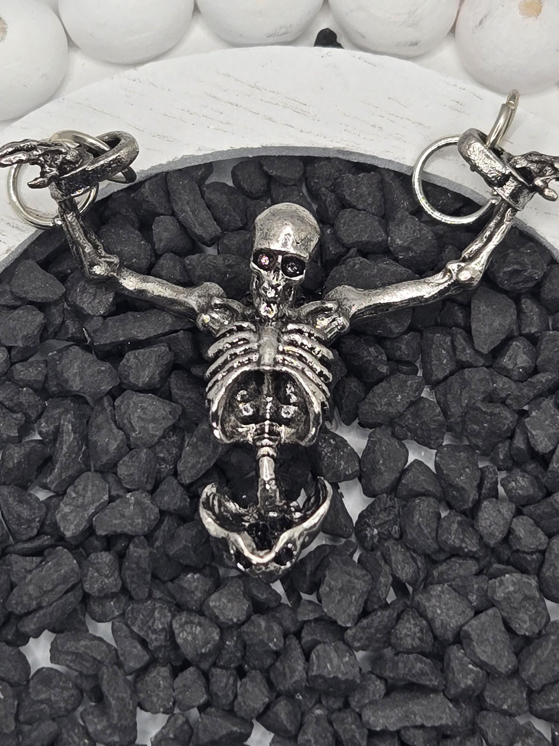 A silver skeleton necklace with a pendant is displayed on a bed of small black stones.