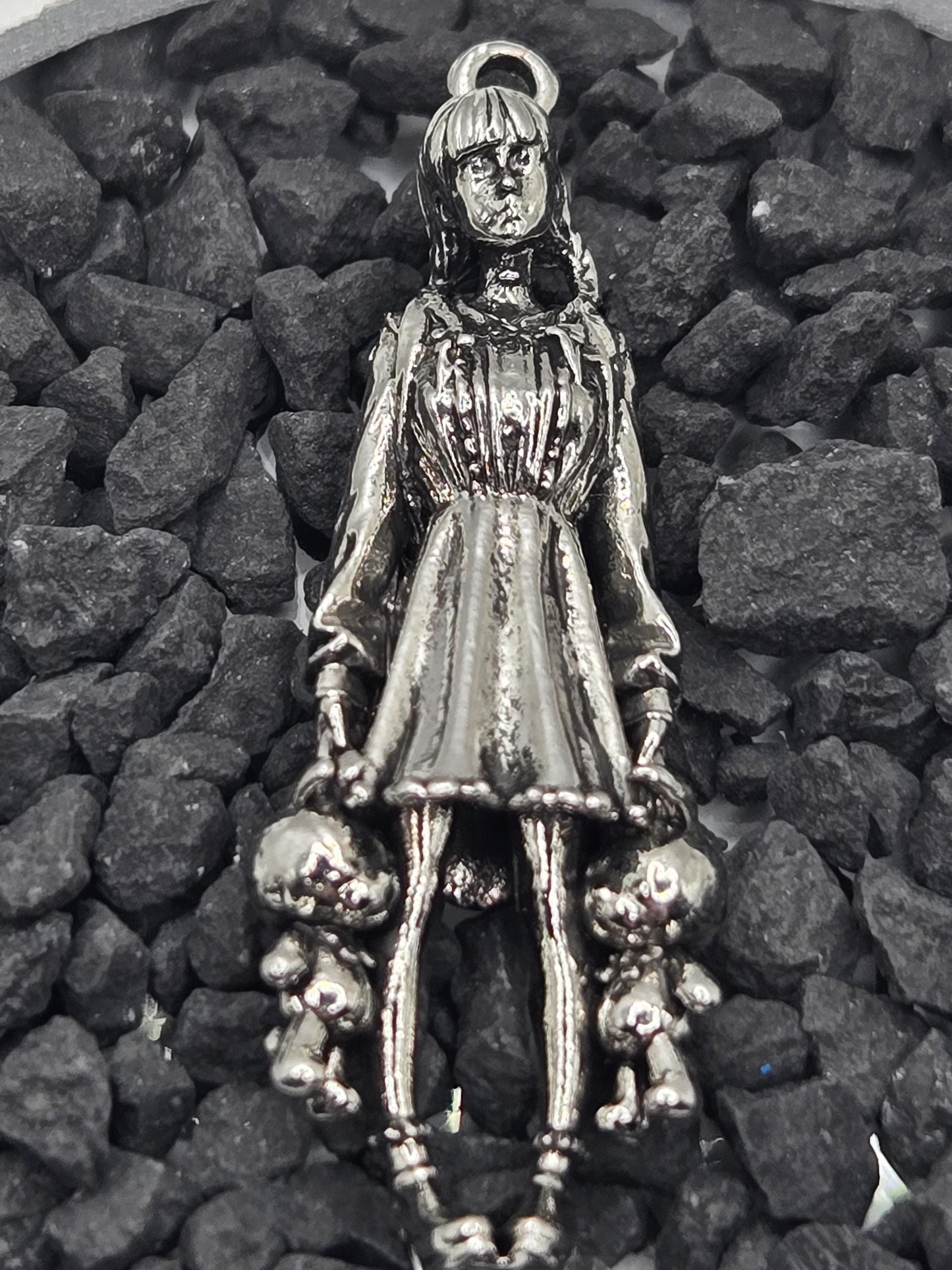 A silver-colored metal charm in the shape of a woman, with a long dress and a headpiece, is resting on a bed of small rocks.